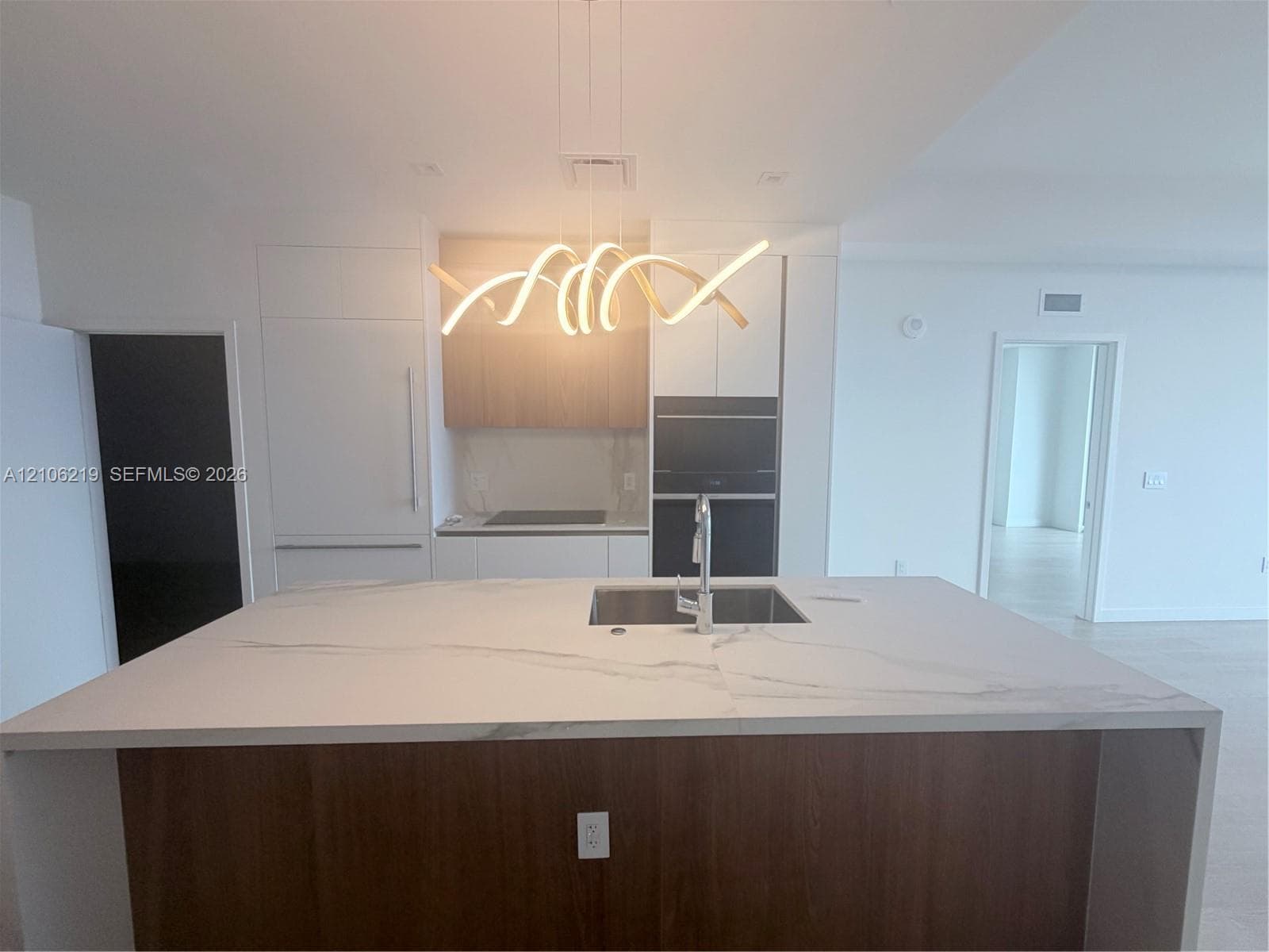 Kitchen featuring a waterfall-edge island with stone-finish countertop, integrated stainless steel sink, custom cabinetry with wood-finish and matte-finish panels, built-in appliances, and a sculptural contemporary chandelier
