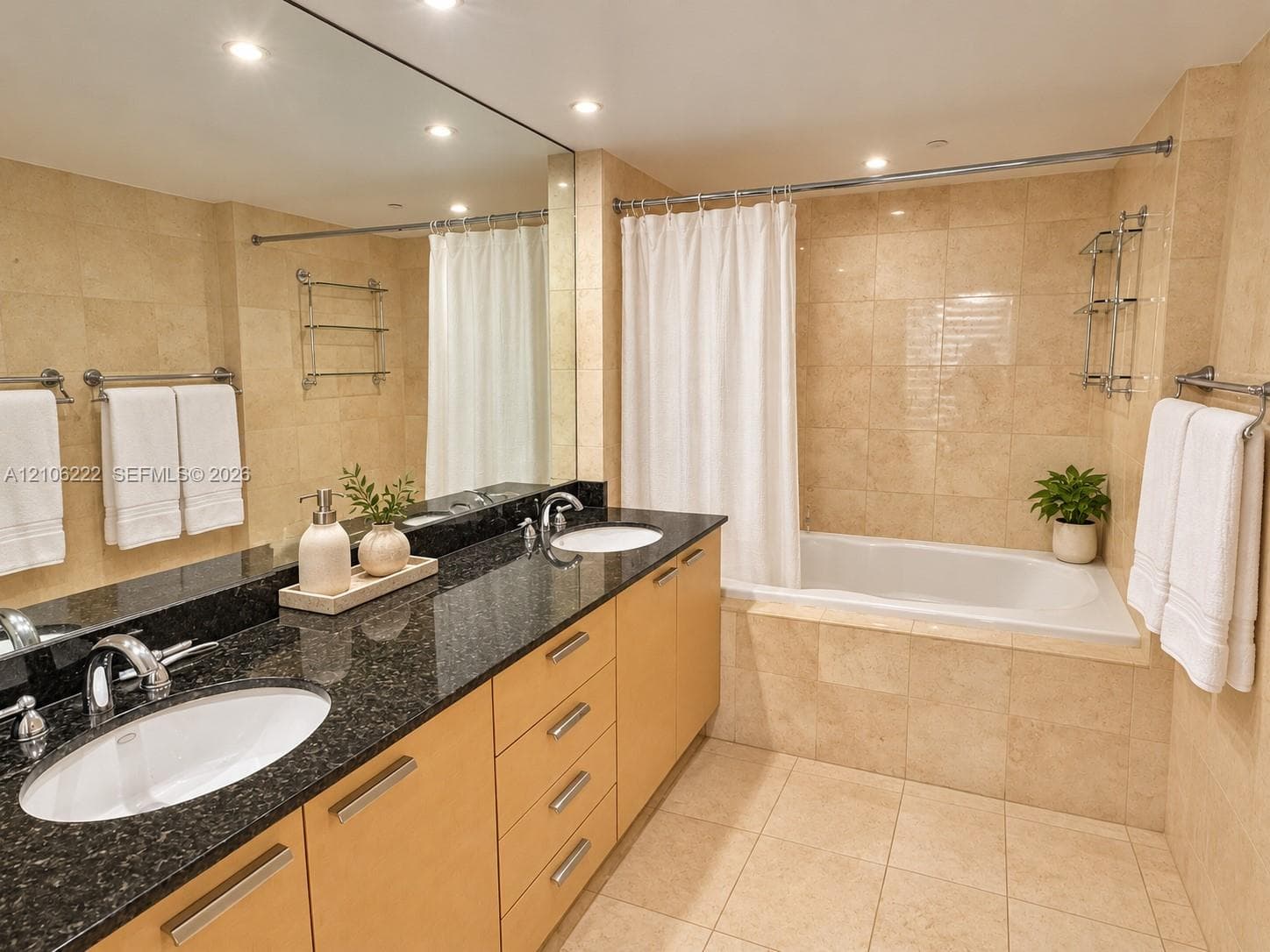 Double vanity with dark stone countertop, integrated dual sinks, light wood-finish cabinetry, floor-to-ceiling tile work, and built-in soaking tub