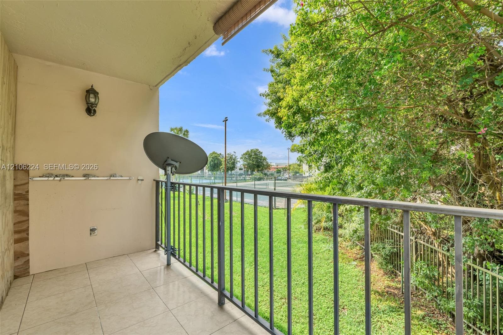 Covered balcony featuring tile flooring, metal railing, wall-mounted exterior light fixture, and mature tree backdrop