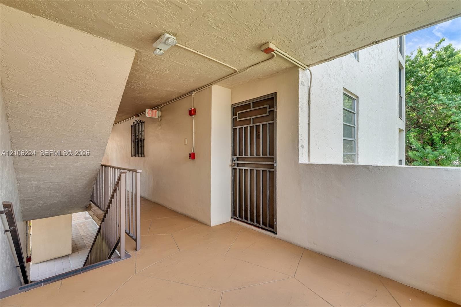 Covered exterior walkway featuring textured stucco walls, tiled flooring, metal security door, and railing-enclosed stairwell