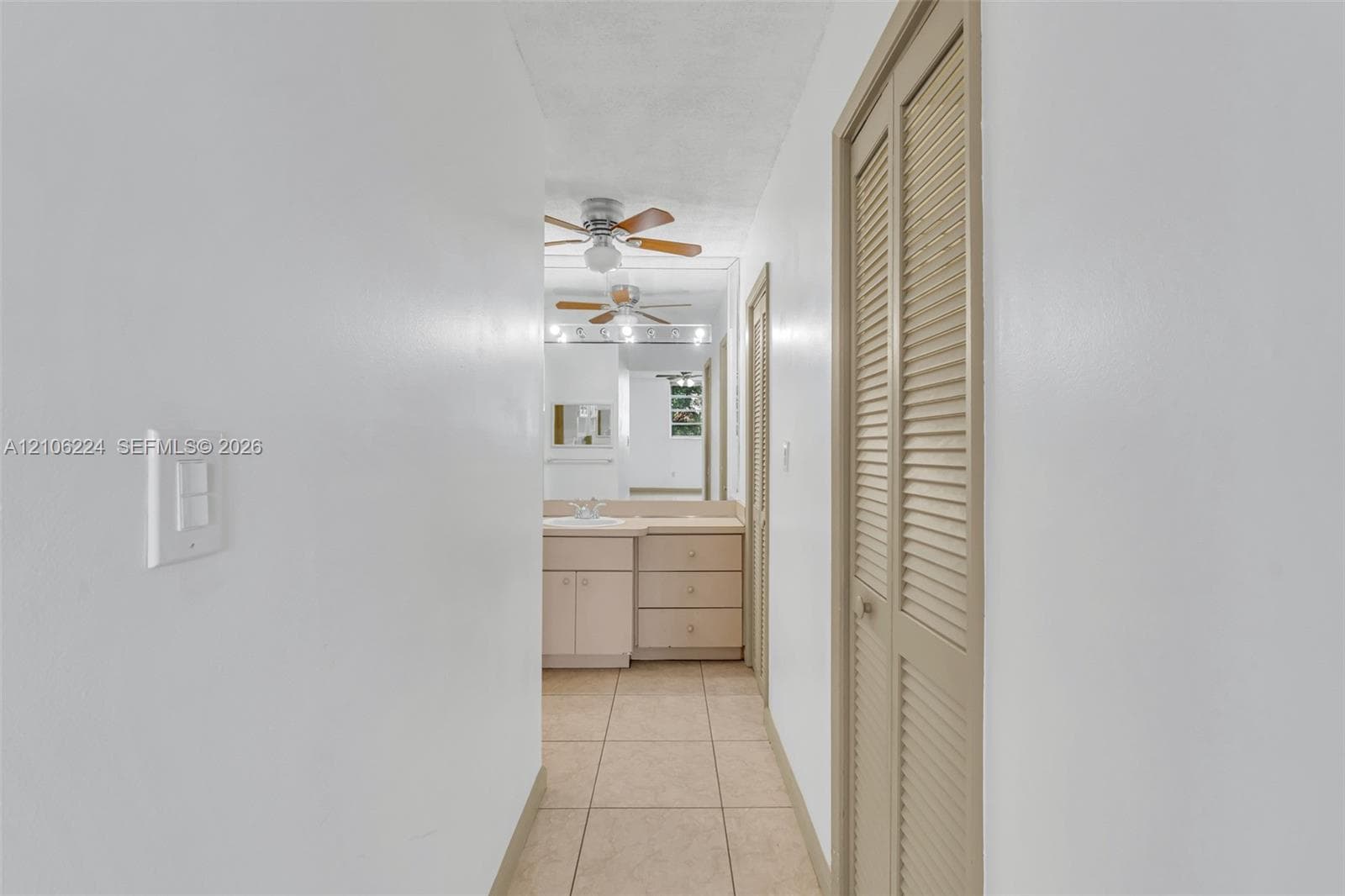 Hallway featuring tile flooring, dual louvered closet doors, and a vanity with integrated cabinetry, capped by a large wall-mounted mirror and ceiling-mounted paddle fan
