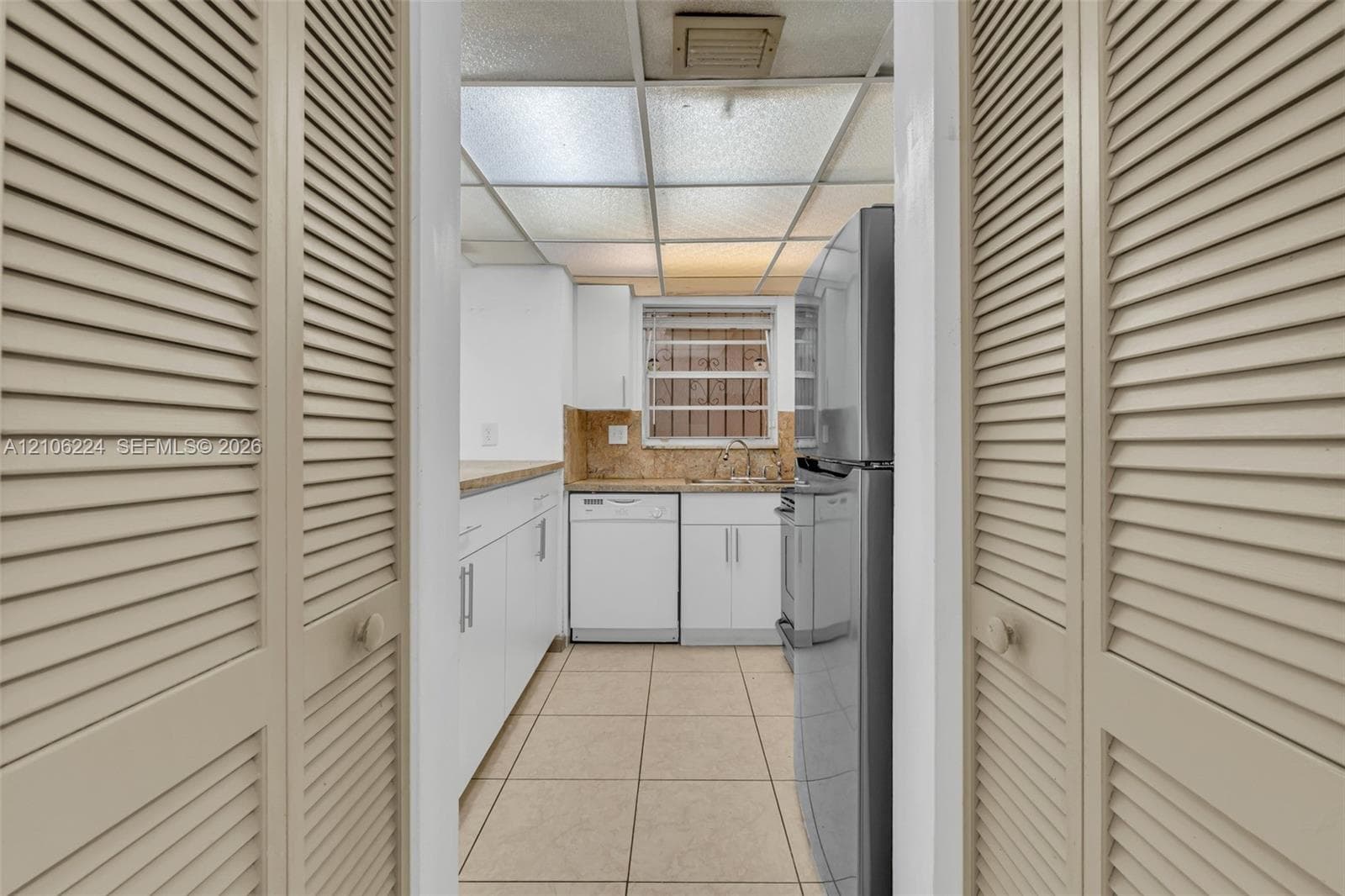 Kitchen area featuring white cabinetry, light-toned stone-finish countertops, stainless steel appliance suite, tile flooring, and louvered closet doors