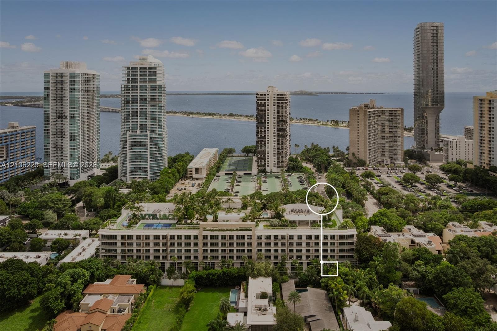 Aerial perspective of a multi-story building complex featuring a rooftop tennis court, mature tree canopy, proximity to the coastline, and surrounding high-rise structures