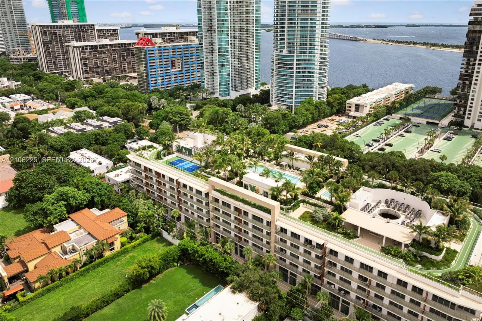 Aerial perspective of a multi-story residential complex featuring a rooftop swimming pool, a blue-surfaced tennis court, a circular courtyard area, and landscaped grounds
