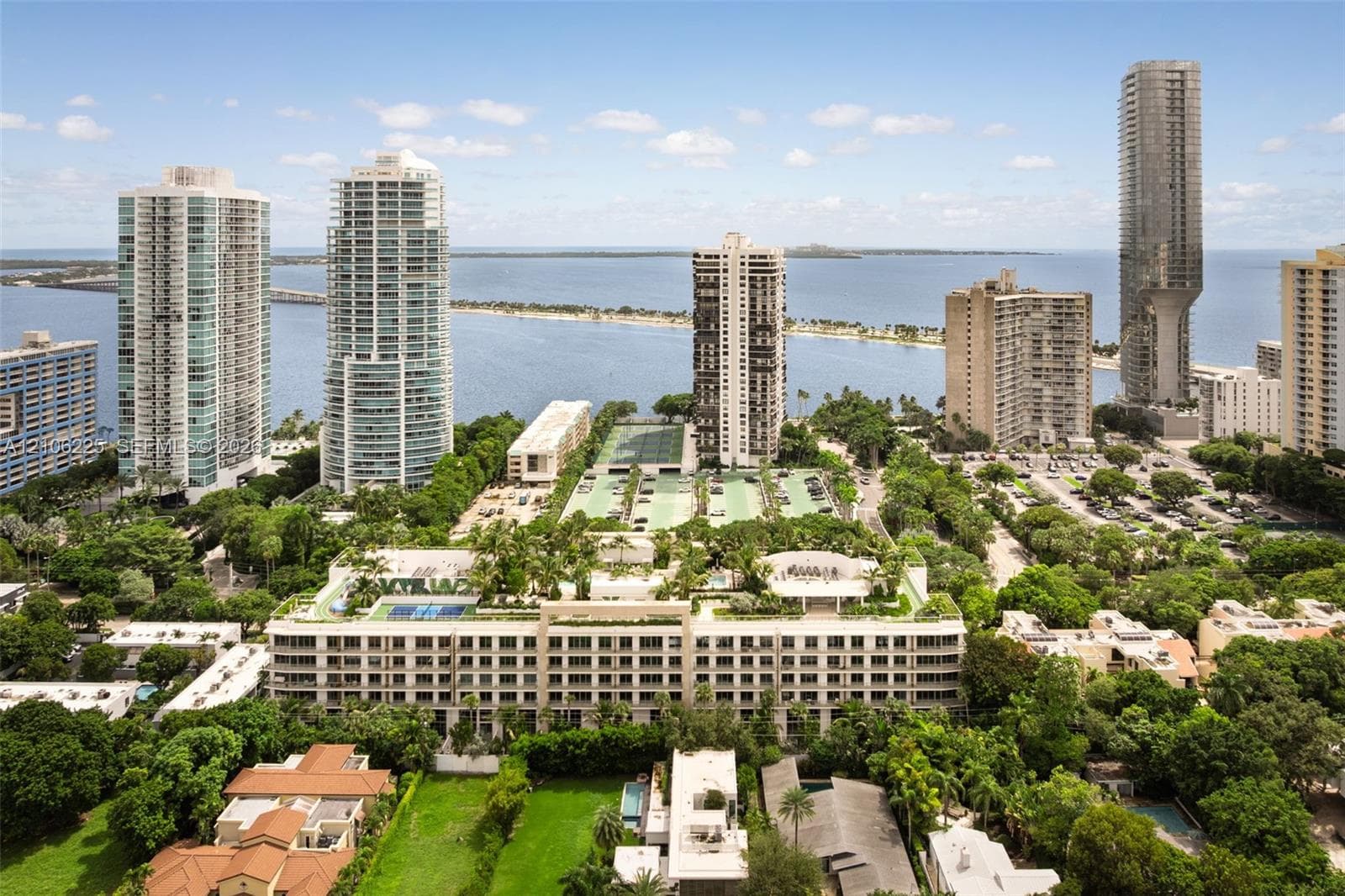 Aerial perspective of a multi-story residential complex featuring a rooftop terrace with multiple swimming pools, professional-grade tennis courts, expansive landscaped grounds, and coastal water proximity