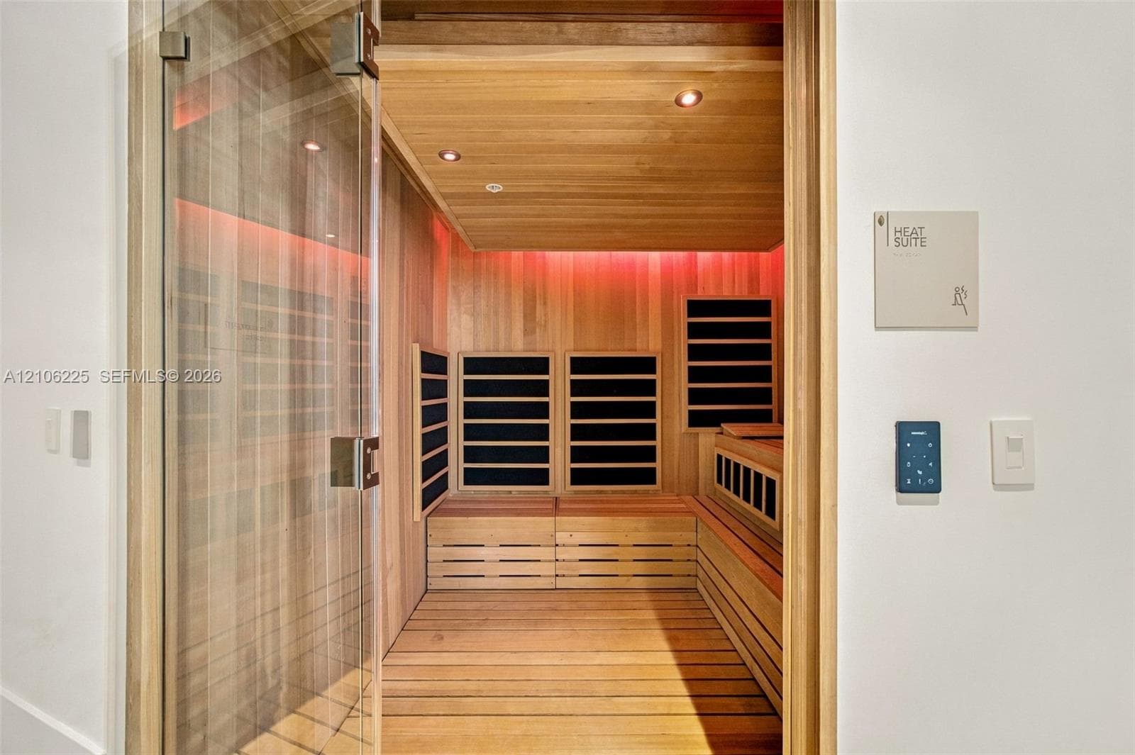 Sauna room featuring light wood-paneled walls and flooring, integrated heating elements, recessed lighting, and a glass door with a polished metallic handle