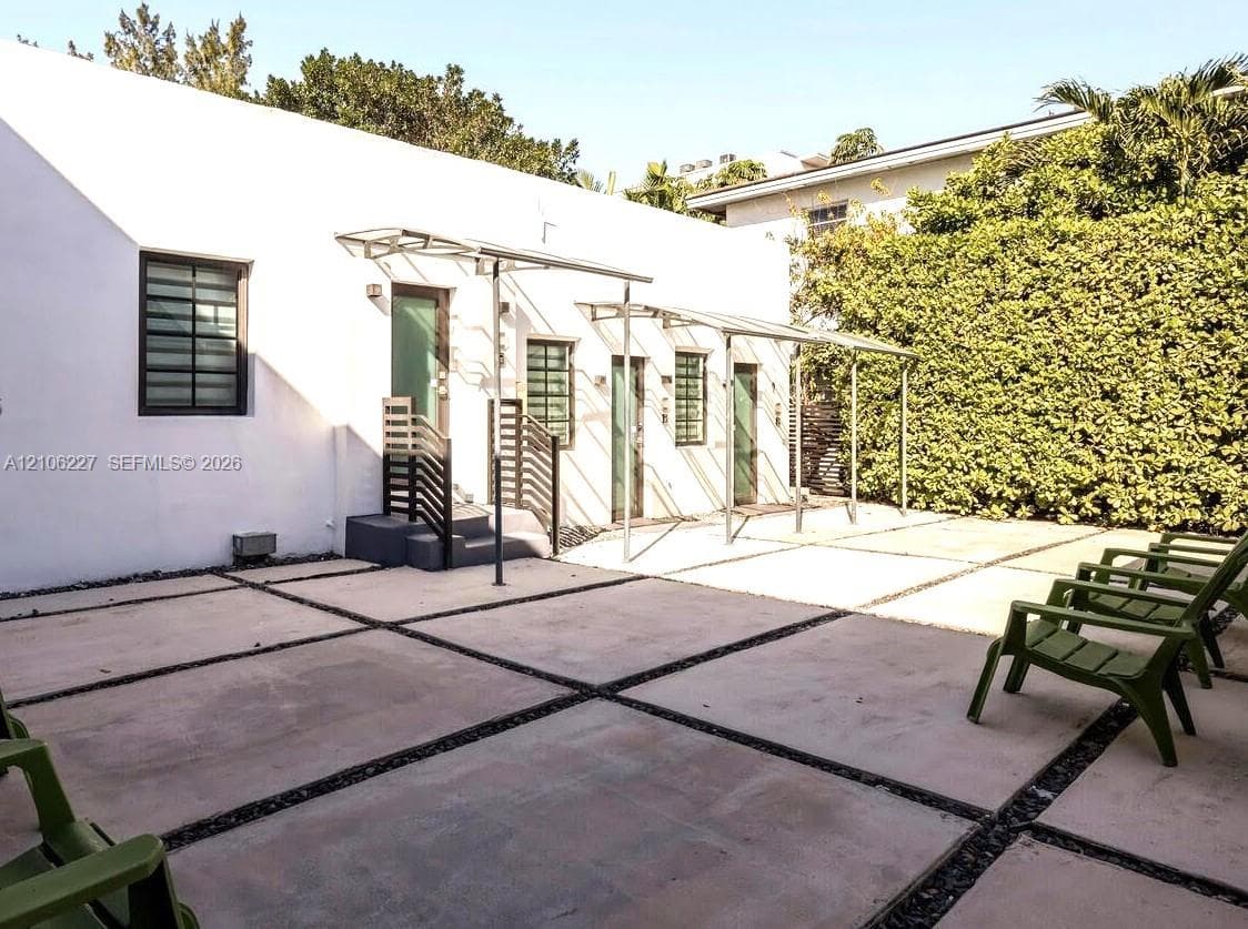 Exterior space featuring a concrete paver patio with gravel detailing, three glass-paneled entry doors, metal awnings, and a privacy hedge