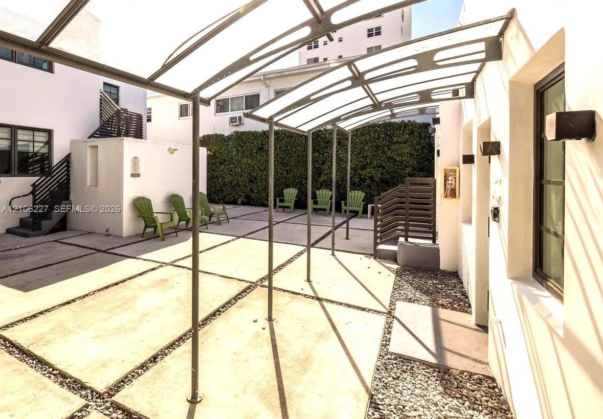 Exterior courtyard featuring a segmented concrete patio with stone-filled borders, a metal-framed overhead canopy, stucco walls, and a tall privacy hedge