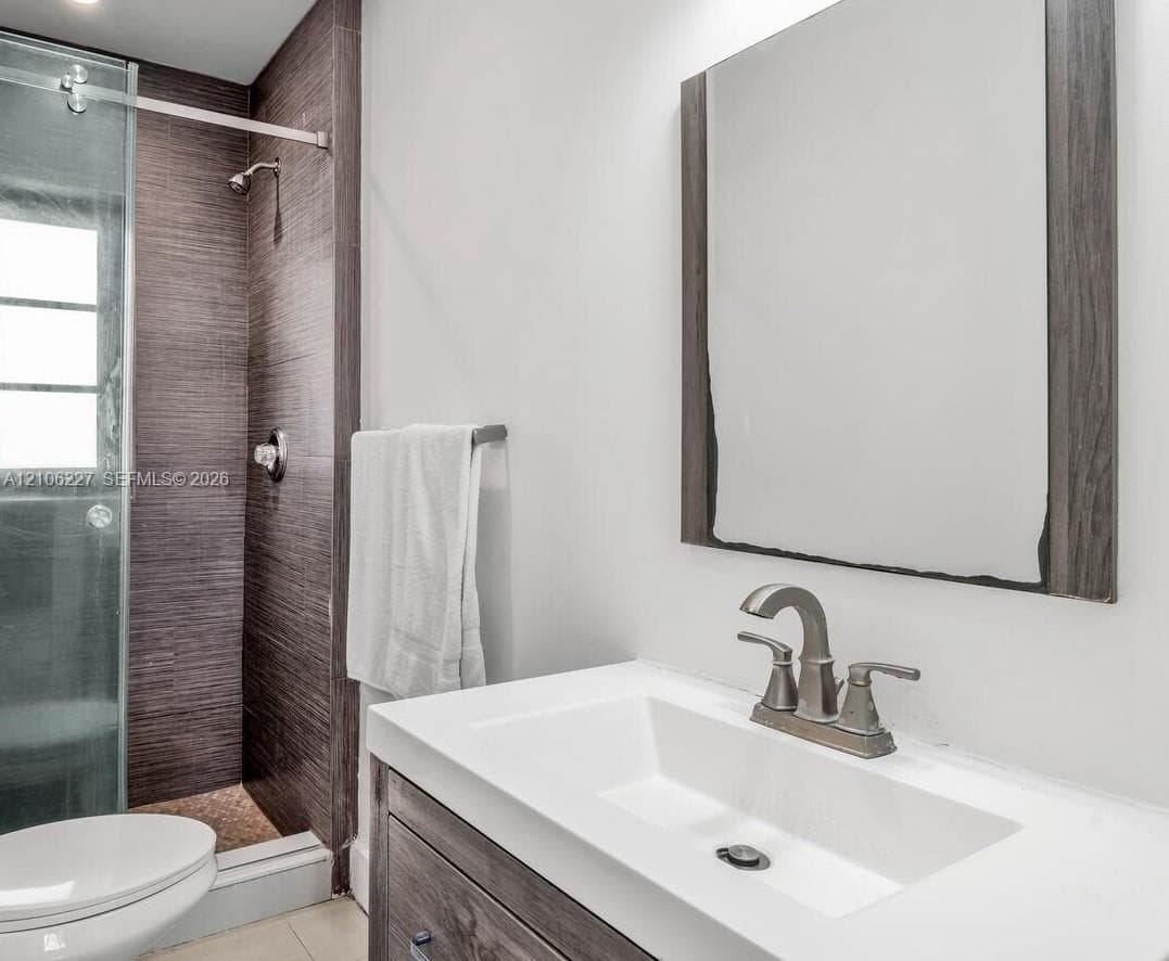 Bathroom featuring a vanity with a white rectangular sink, metallic faucet, wood-finish cabinetry, a framed mirror, and a glass-enclosed shower with textured wall tile