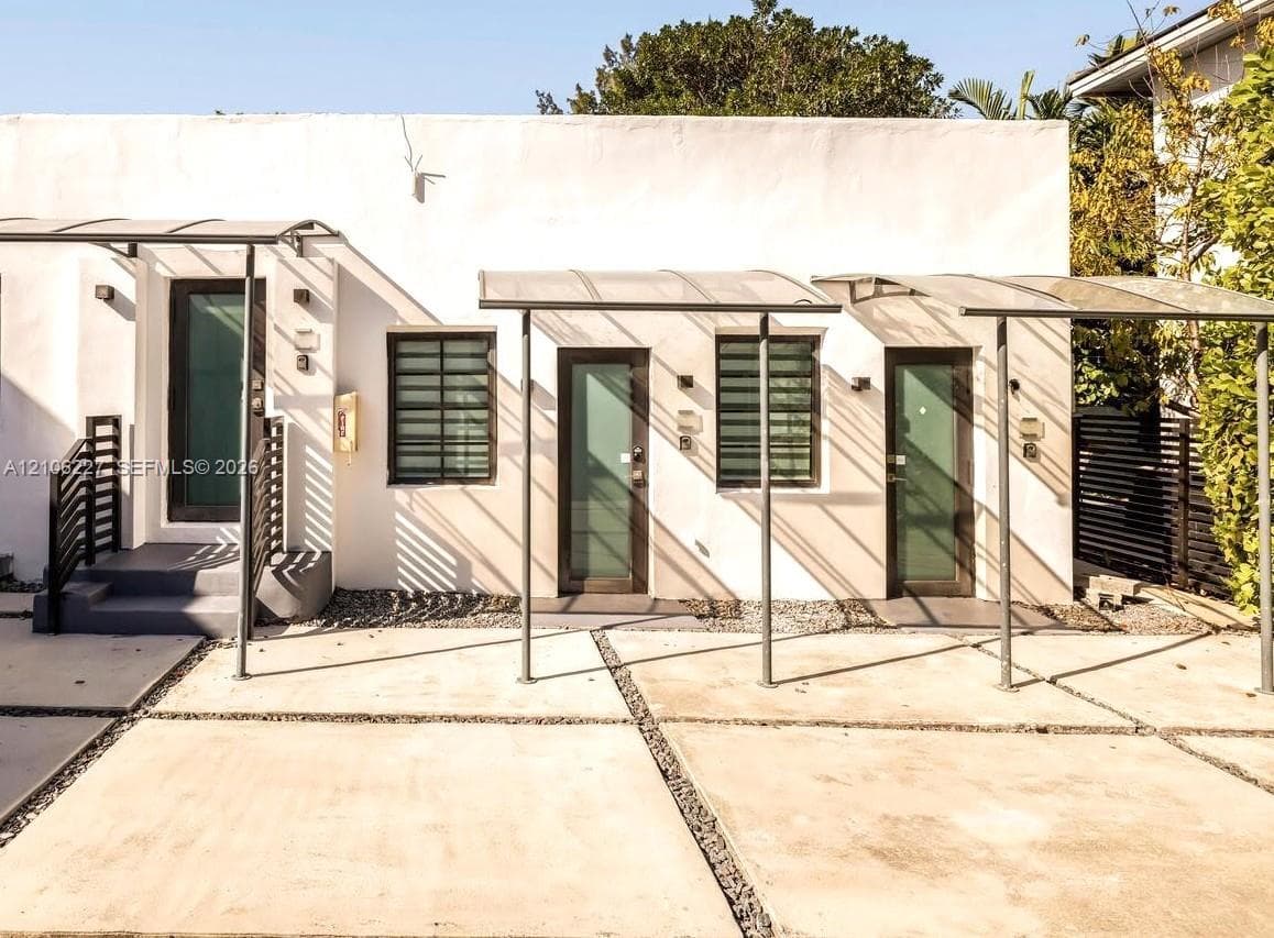 Stucco exterior featuring three glass-panel entry doors, aluminum frame awnings, louvered windows, and a paved concrete parking pad with gravel drainage channels