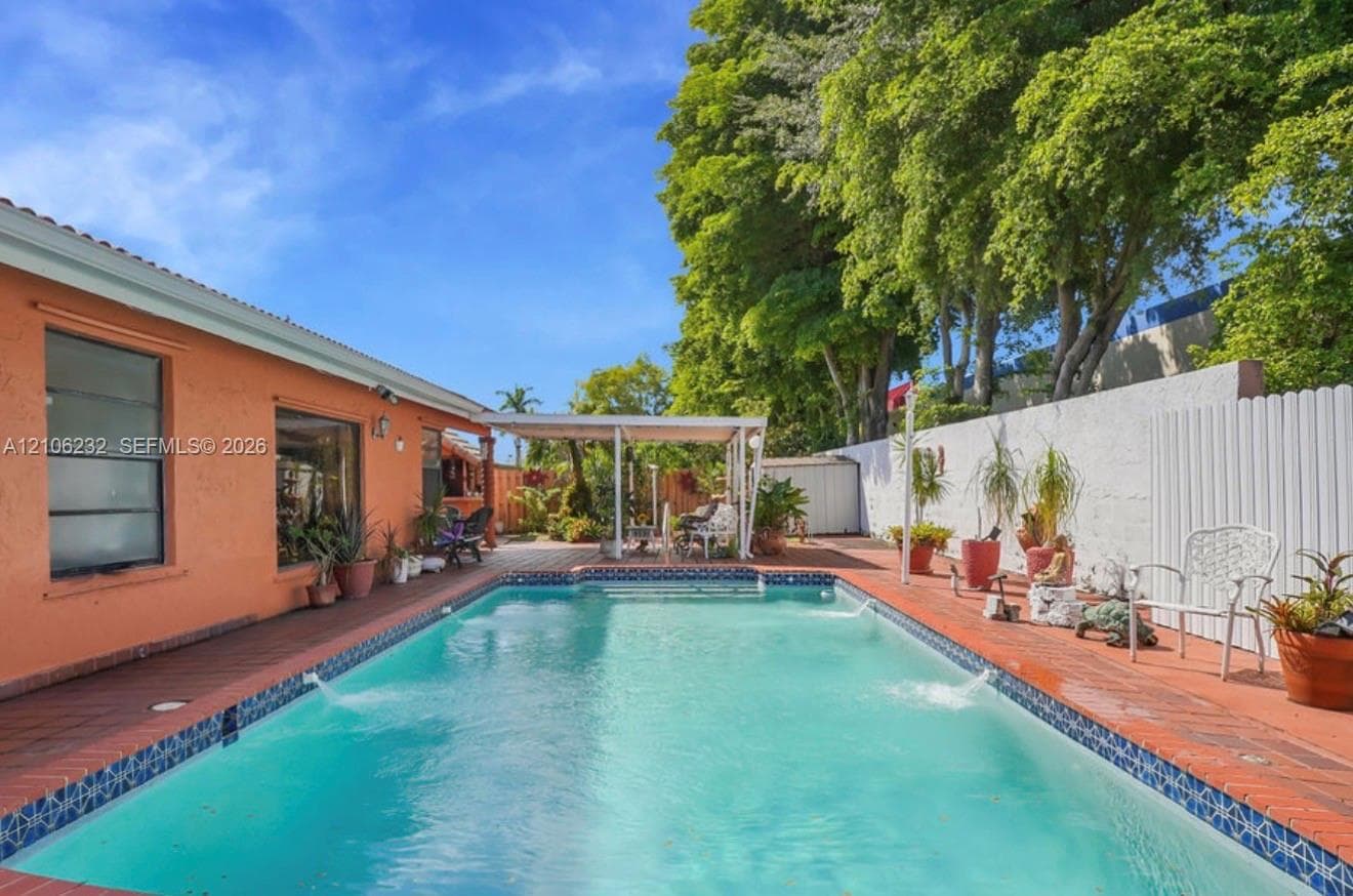 Outdoor pool with decorative tile border and built-in water features, surrounded by a paved brick patio, covered pergola, and privacy wall