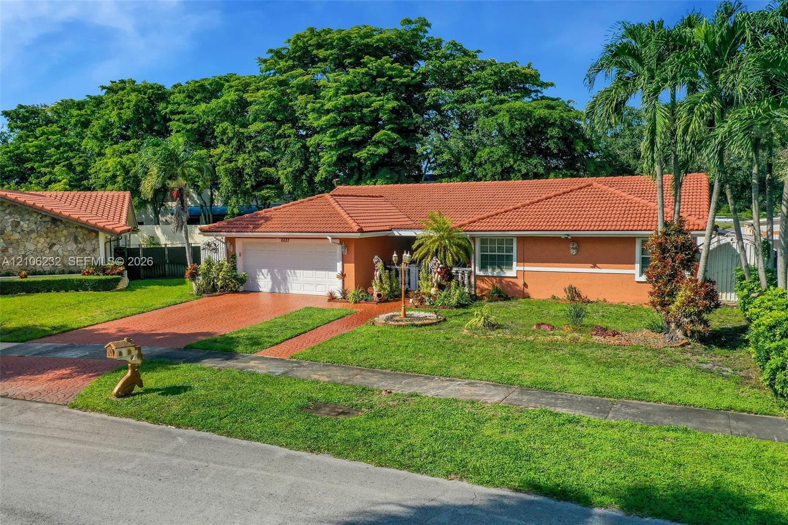 Single-story exterior featuring a terracotta-tiled roof, stucco facade, paved brick driveway, and established landscape including mature palm trees
