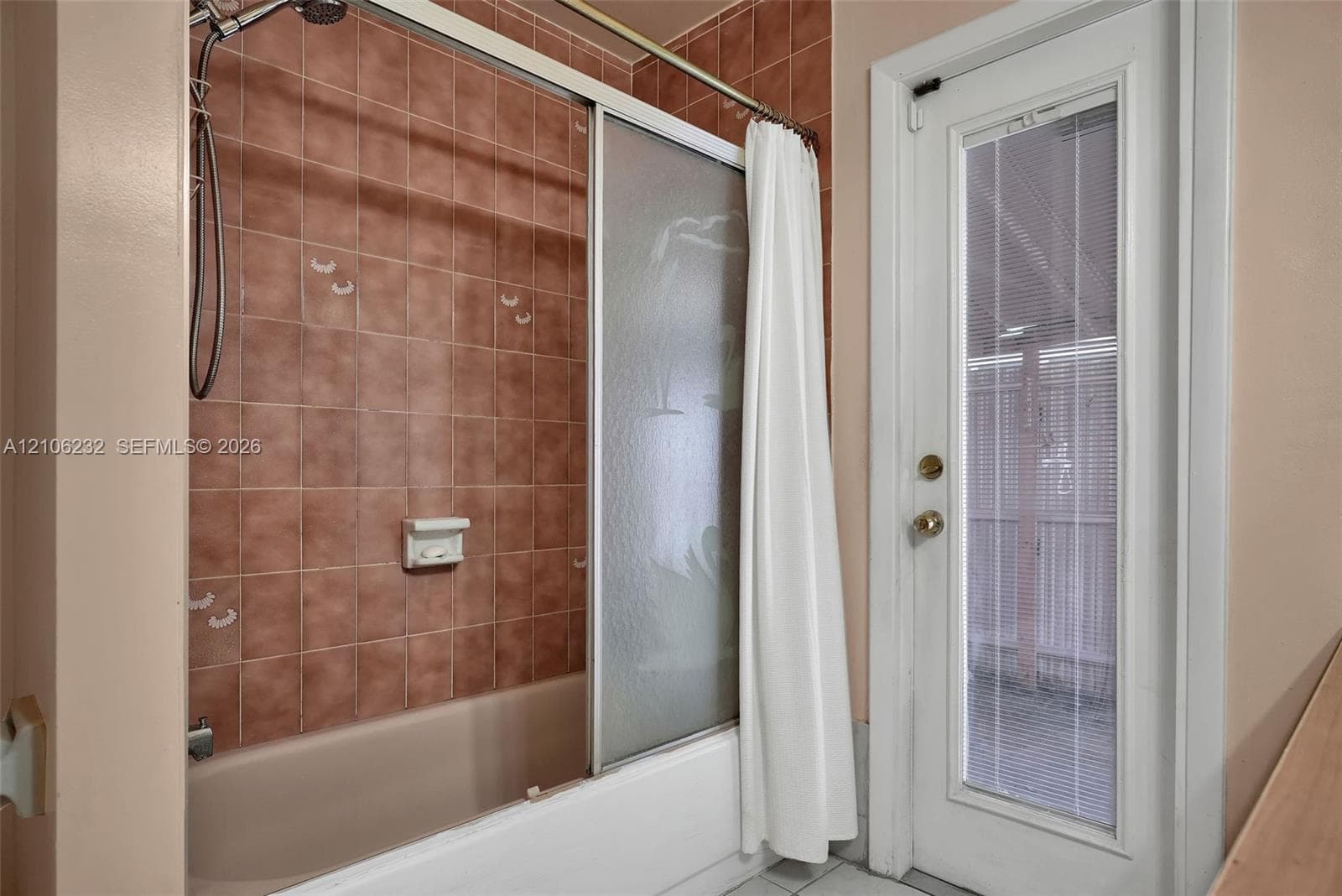 Bathtub with a handheld shower fixture, sliding frosted glass doors, and tiled surround, featuring a full-length exterior door with integrated blinds