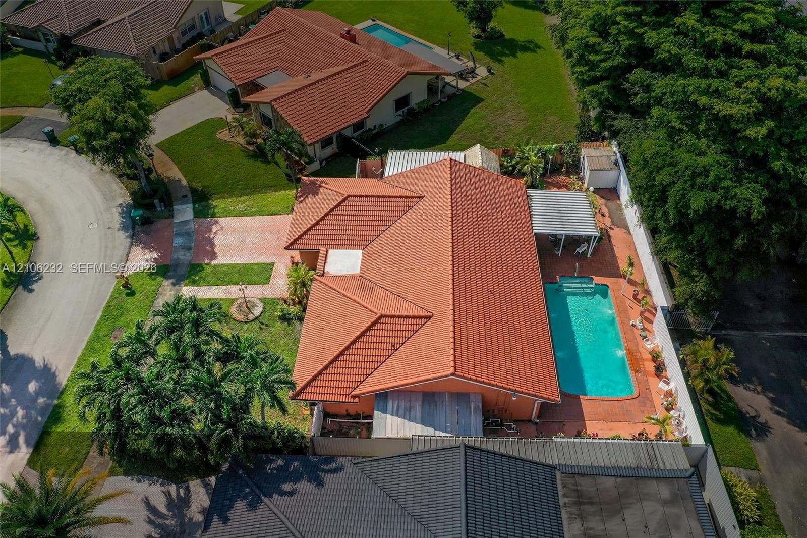 Aerial perspective of a single-story residence featuring a terracotta-tiled roof, an in-ground swimming pool, a brick-paved patio, a metal-framed pergola, and mature landscaping