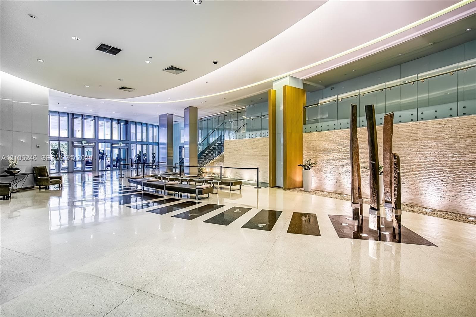 Grand lobby featuring polished stone flooring, curved recessed ceiling lighting, full-height floor-to-ceiling windows, stacked-stone accent wall, and glass-paneled staircase railing