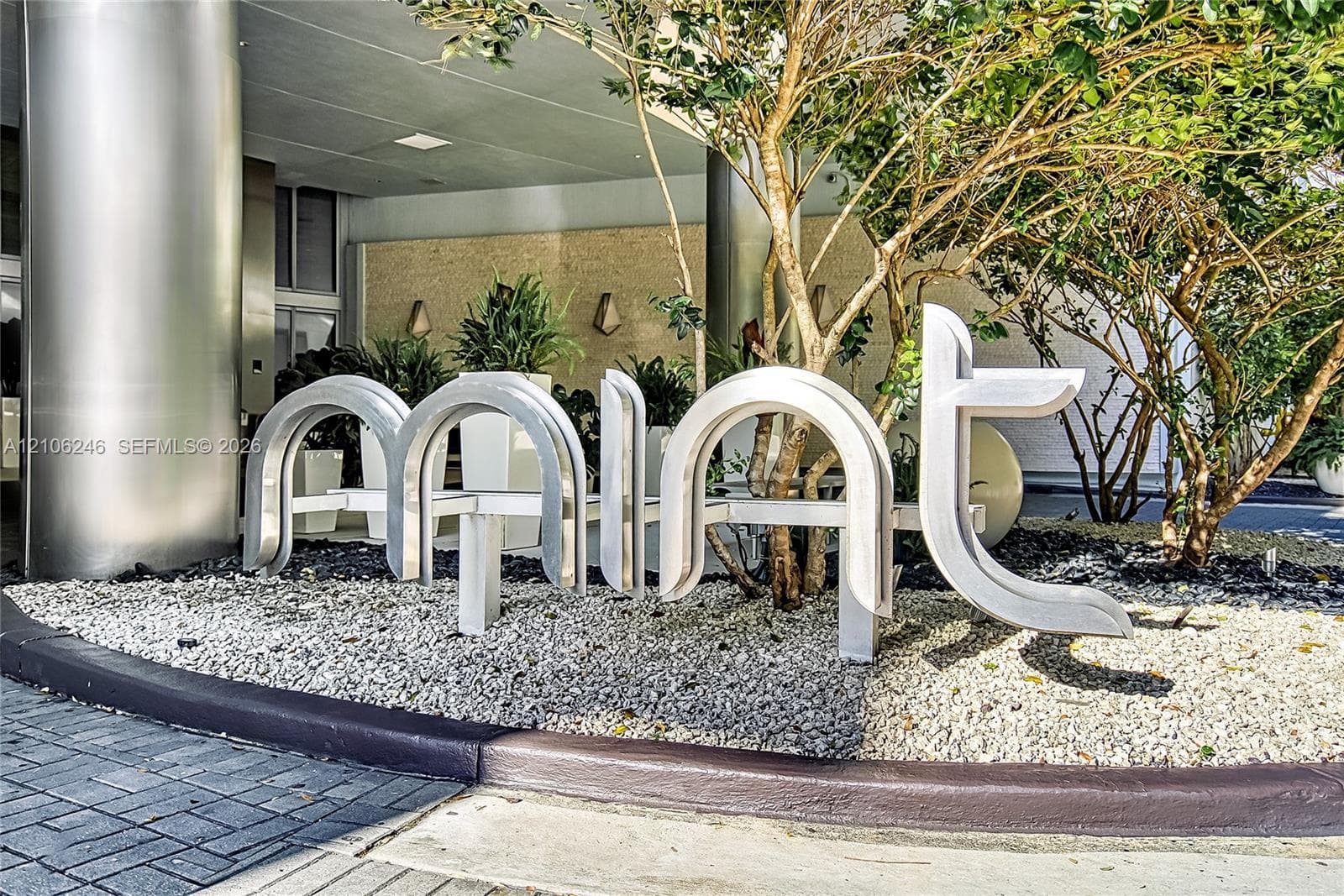 Exterior entrance featuring brushed metal structural columns, stone-textured facade wall, sculptural metal signage, and gravel landscaping with integrated greenery