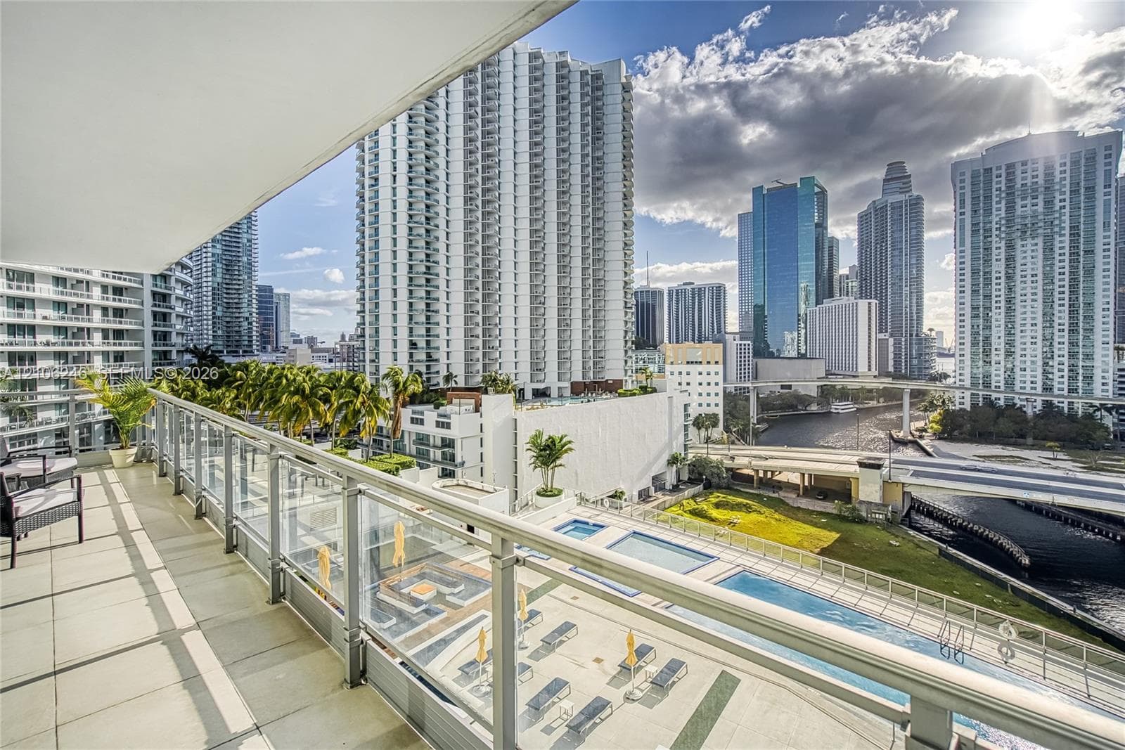 Covered balcony featuring tile flooring and a glass-paneled railing overlooking an urban skyline and multi-level swimming pools