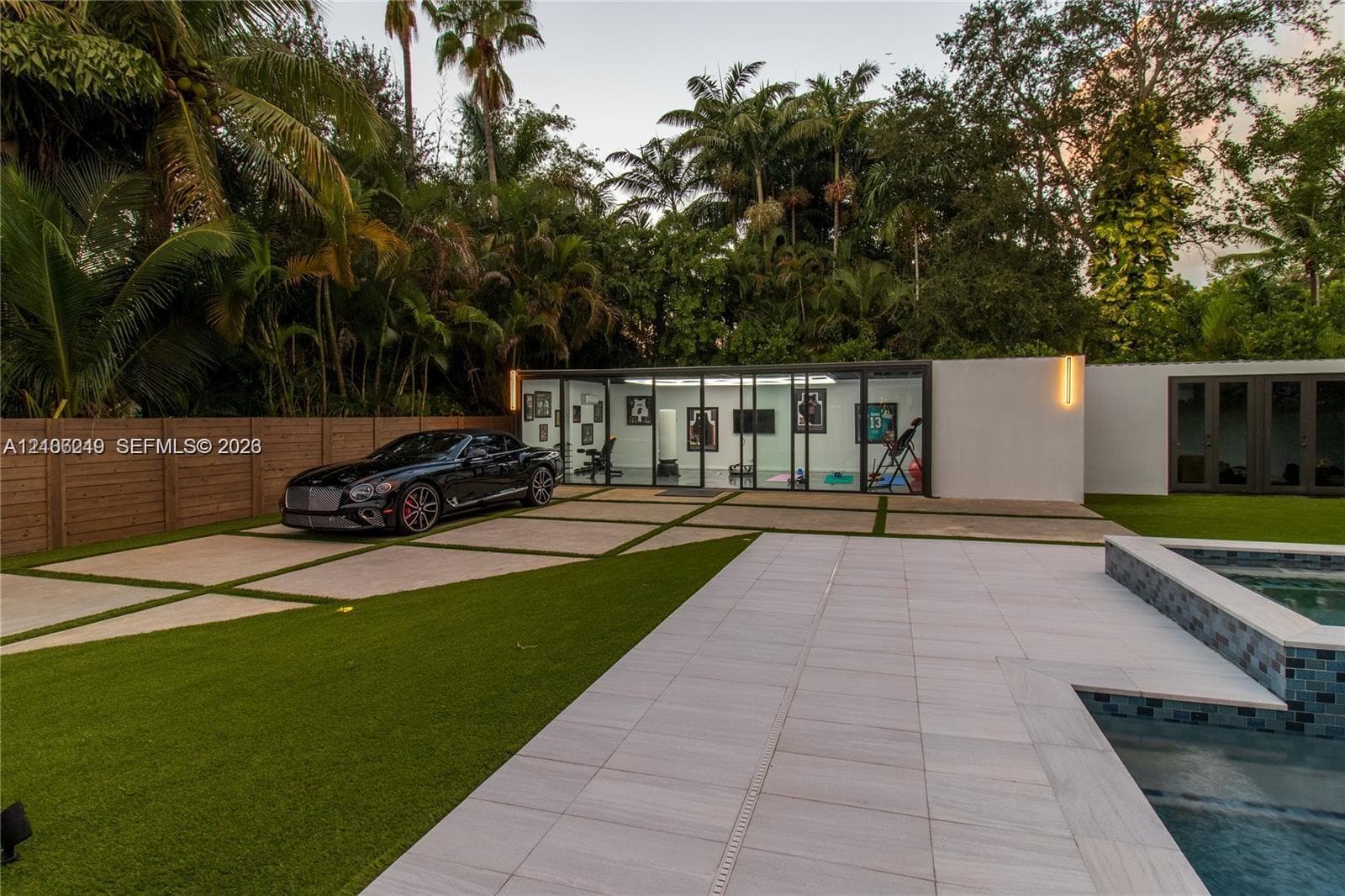 Contemporary pool deck featuring large-format stone pavers, integrated glass-walled studio structure, geometric concrete driveway with turf inserts, and perimeter wood privacy fencing