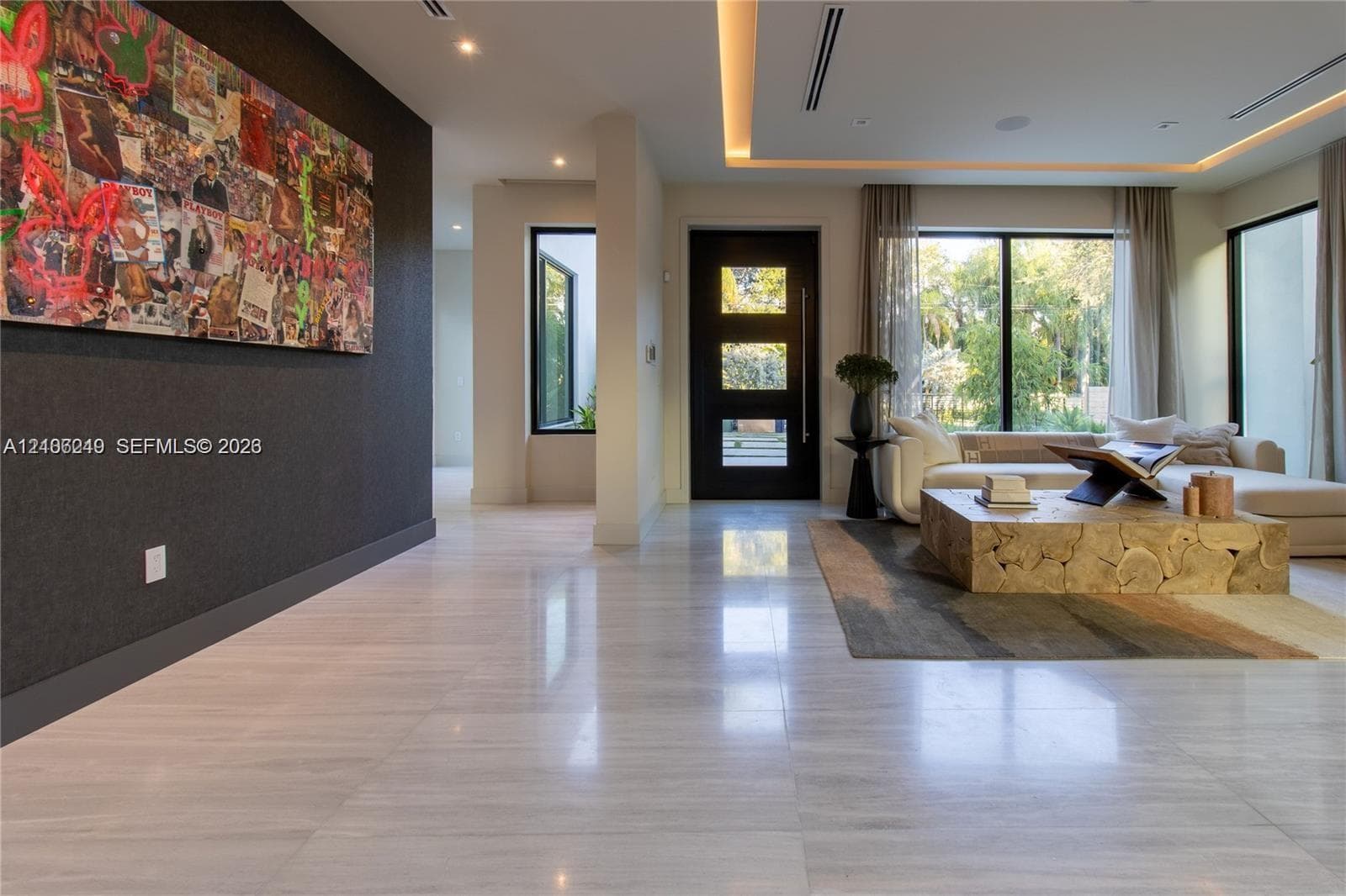 Large-format polished stone flooring, recessed tray ceiling with integrated cove lighting, dark solid-core entry door with glass inserts, and floor-to-ceiling windows