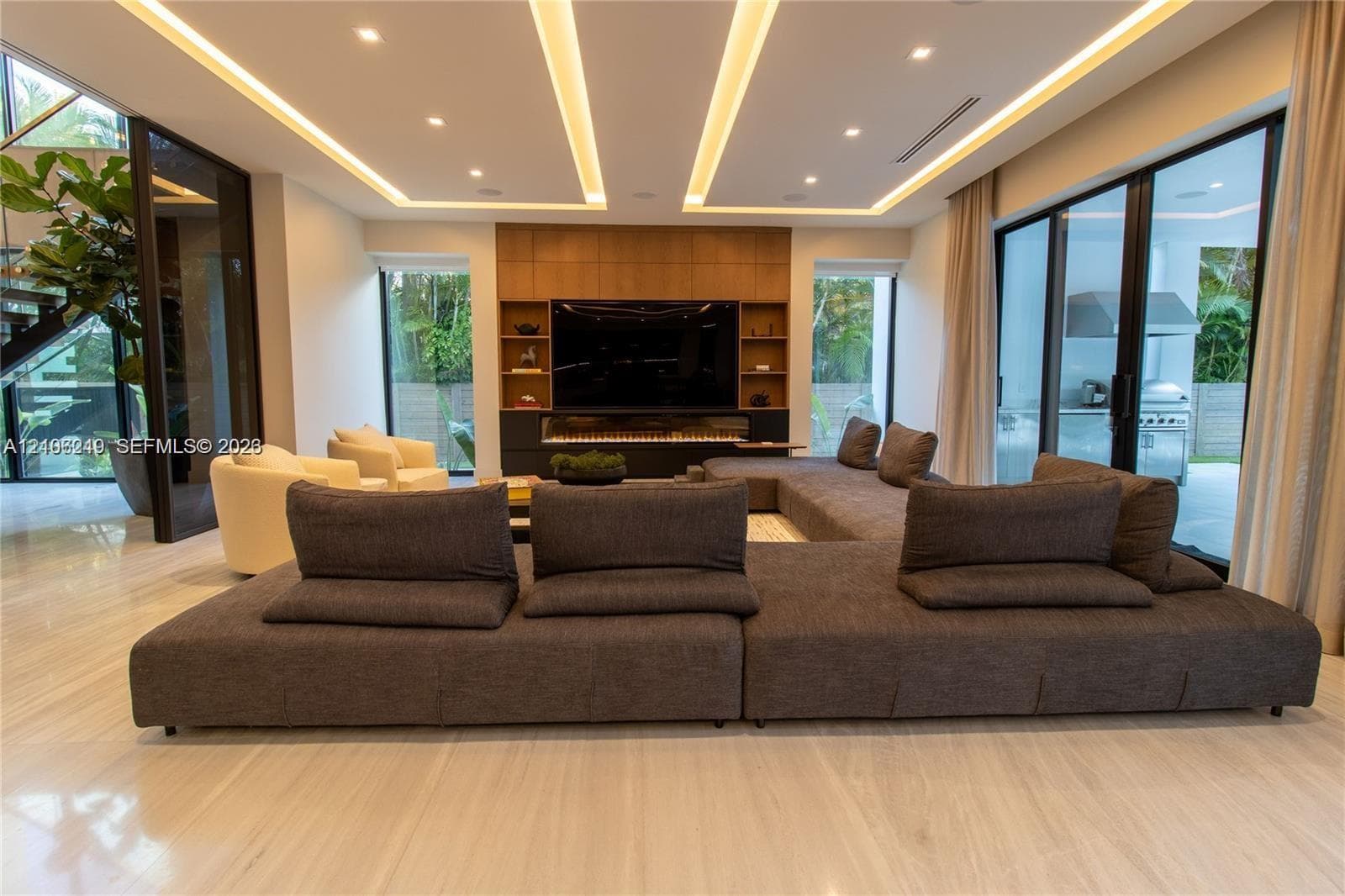 Living area featuring a coffered ceiling with integrated cove lighting, recessed lighting, polished stone flooring, a full-height wood-paneled media wall with built-in shelving, and floor-to-ceiling glass sliding doors