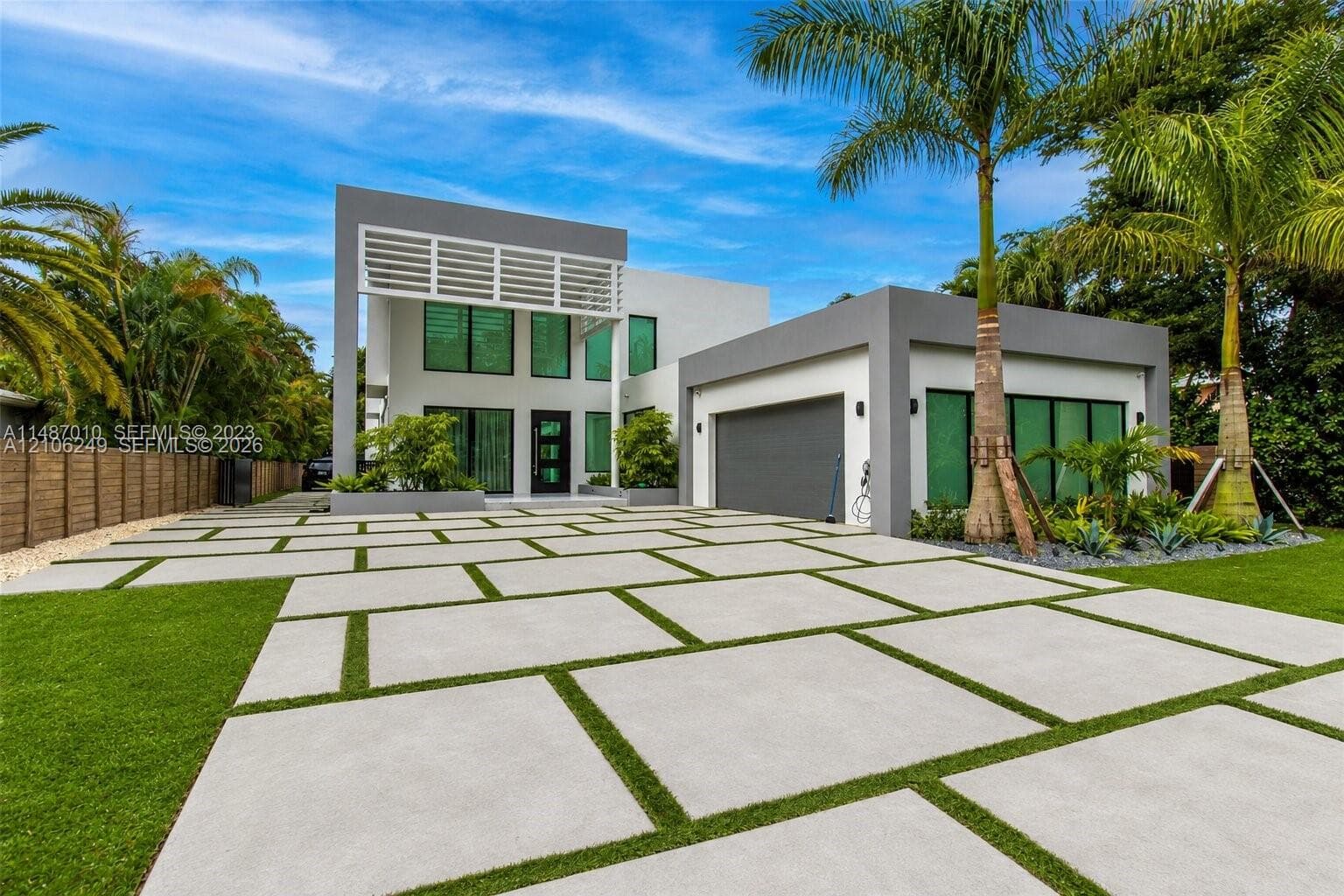 Modern two-story exterior featuring a geometric concrete paver driveway, white slatted architectural detail, expansive floor-to-ceiling windows, and integrated garage