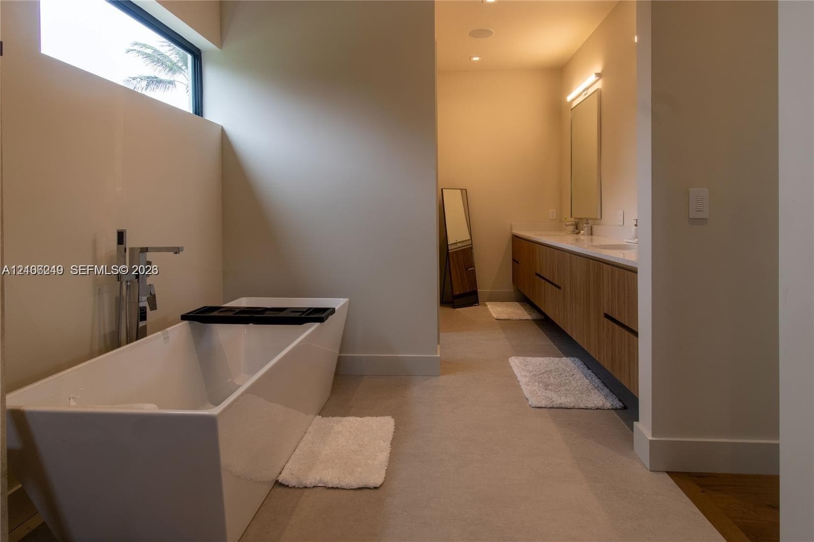 Freestanding soaking tub with floor-mounted faucet, horizontal clerestory window, dual-sink vanity with wood-finish cabinetry, and neutral large-format tile flooring