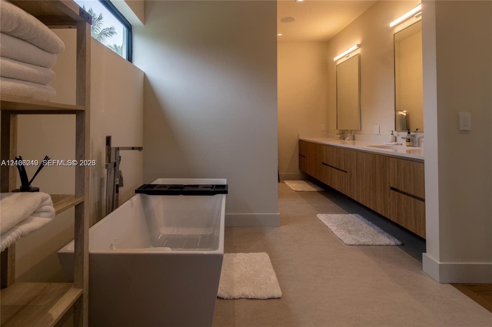 Freestanding soaking tub with floor-mounted faucet, dual-sink floating vanity with wood-finish cabinetry, countertop-to-ceiling mirrors, integrated wall lighting, and horizontal transom window