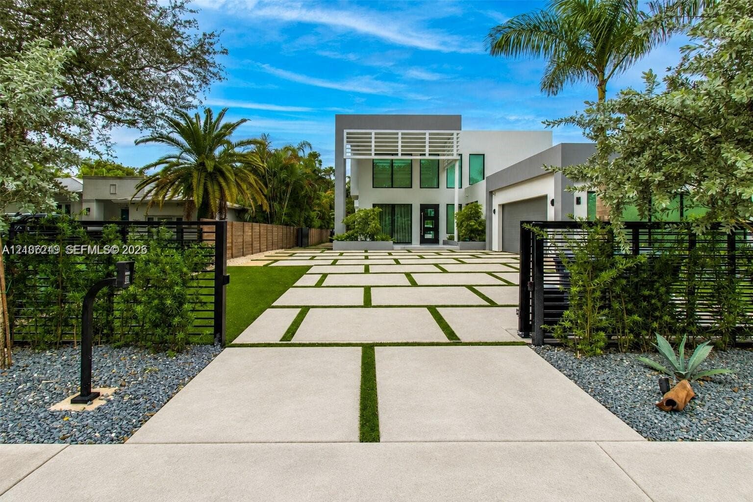 Contemporary residence featuring a geometric concrete driveway with turf ribbons, vertical louvered architectural accents, full-height windows, a metal-framed privacy gate, and a curated landscape with palms