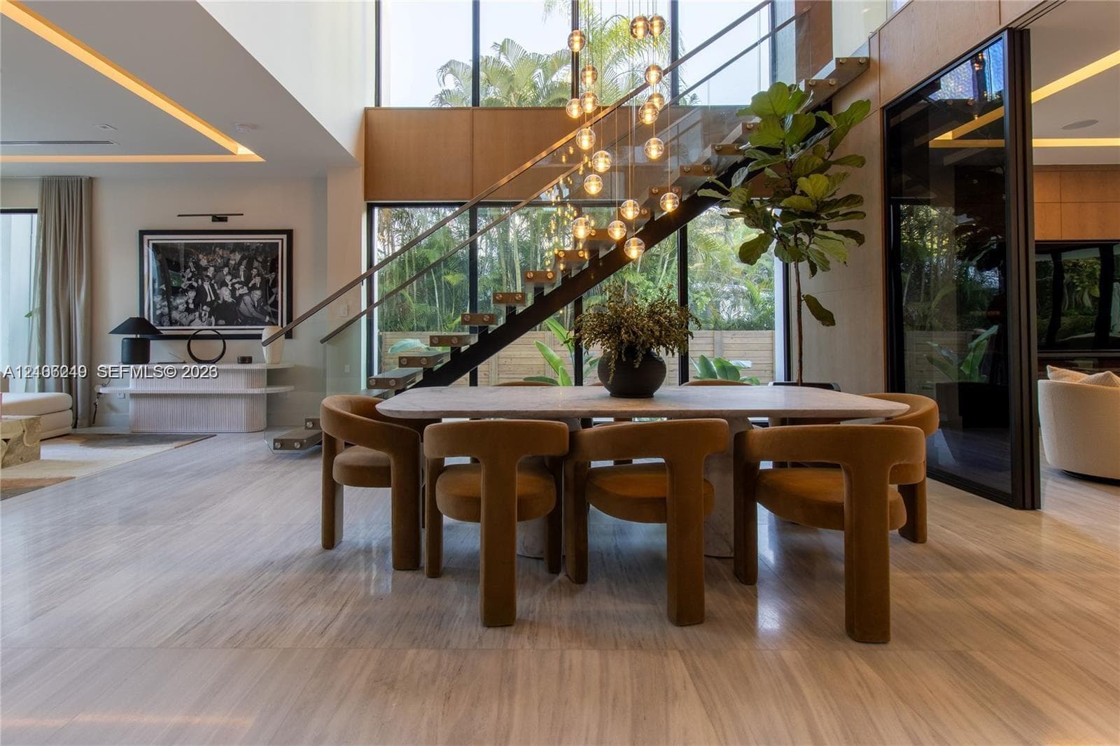 Double-height ceiling featuring floor-to-ceiling windows, open-riser staircase with glass balustrade, stone flooring, integrated recessed lighting, and multi-pendant chandelier