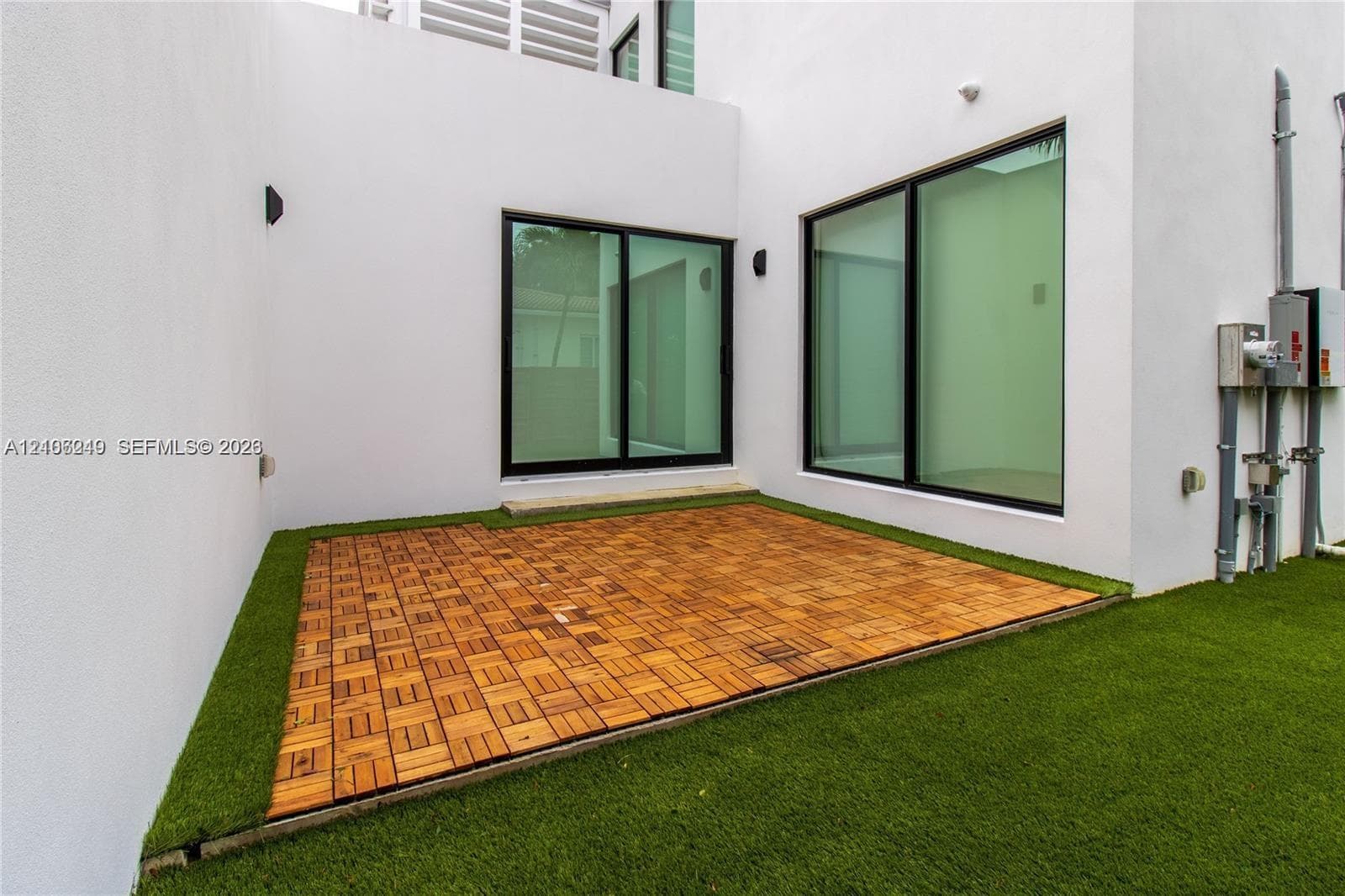 Modern outdoor patio featuring modular wood-finish decking, synthetic turf, minimalist white stucco exterior walls, and floor-to-ceiling sliding glass doors with black frames