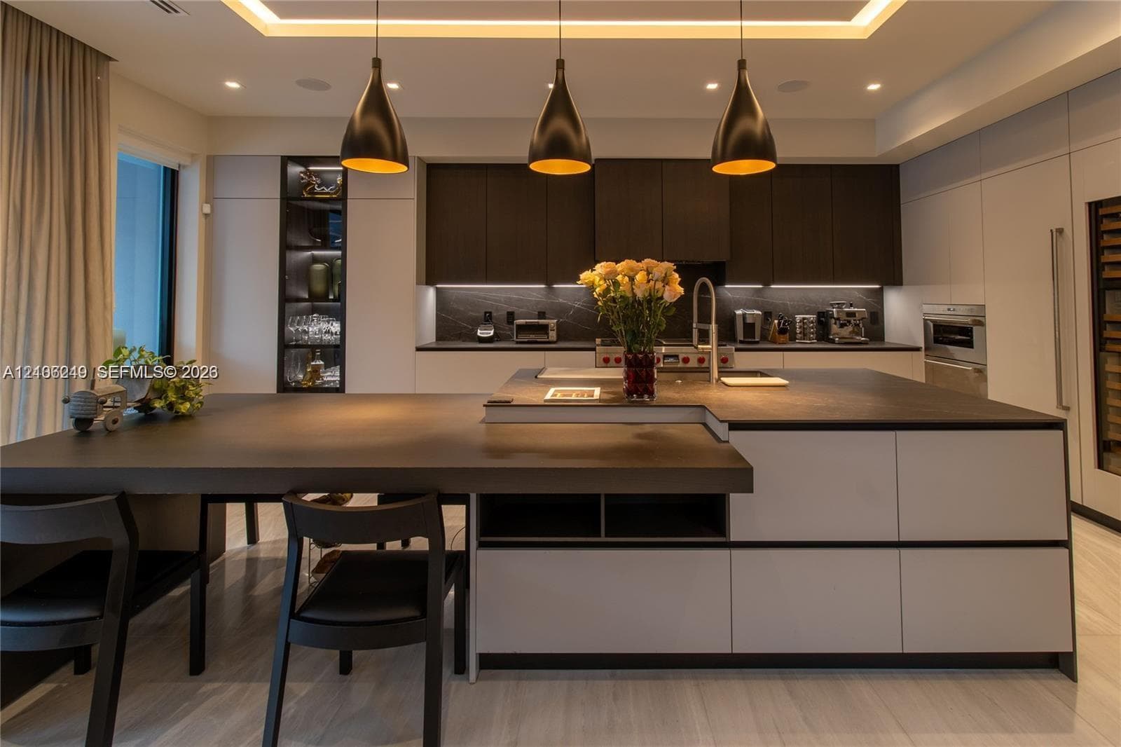 Modern kitchen featuring dark-toned cabinetry, a substantial kitchen island with an integrated dining surface, recessed ceiling lighting, three suspended pendant lights, and built-in appliances