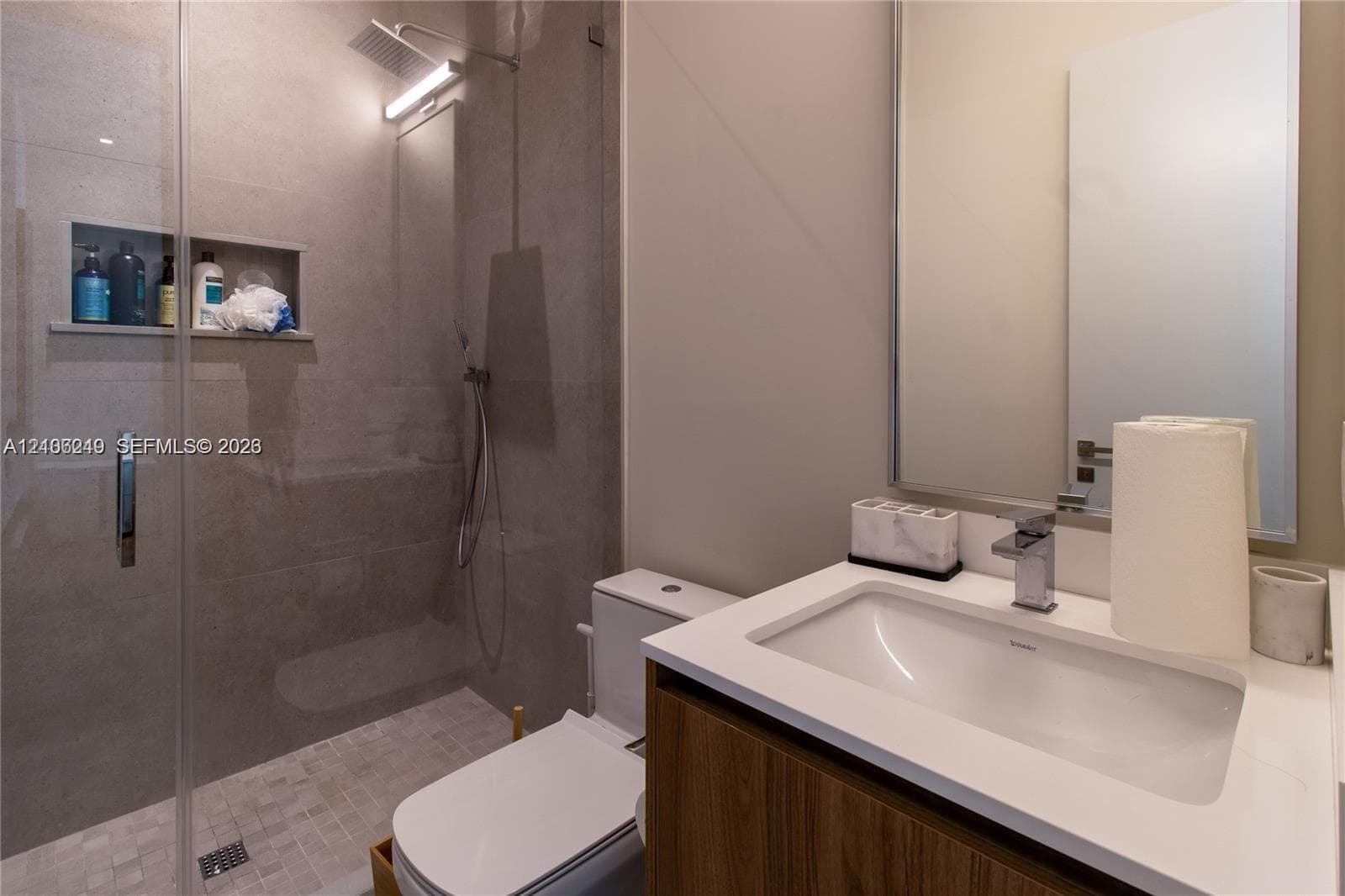 Bathroom featuring a glass-enclosed walk-in shower with stone tile, a built-in recessed niche, rainfall showerhead, wood-finish vanity with an integrated sink, and a quartz countertop