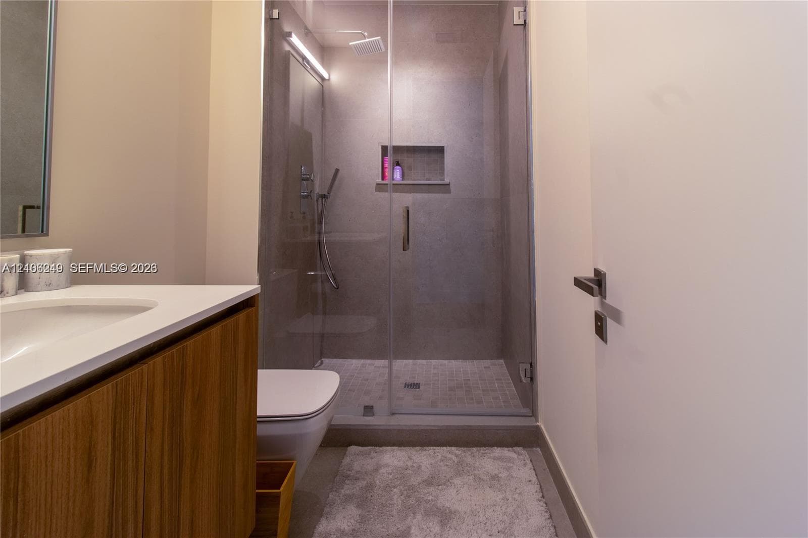Wood-finish vanity cabinet with a white countertop and integrated sink, glass-enclosed shower featuring tiled walls, built-in recessed shelving, a rain-style showerhead, and a wall-mounted toilet