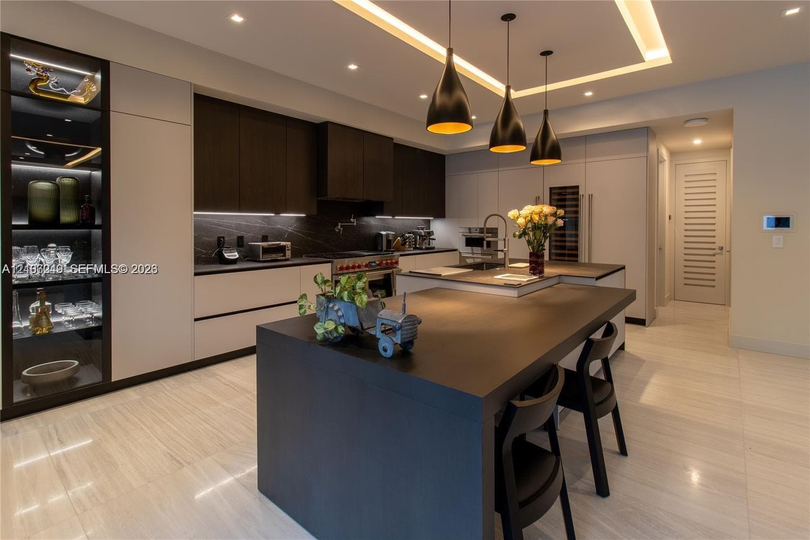 Gourmet kitchen featuring flat-panel cabinetry, a central waterfall-edge island, stone-surfaced countertops, custom recessed tray ceiling lighting, and integrated glass-front display shelving