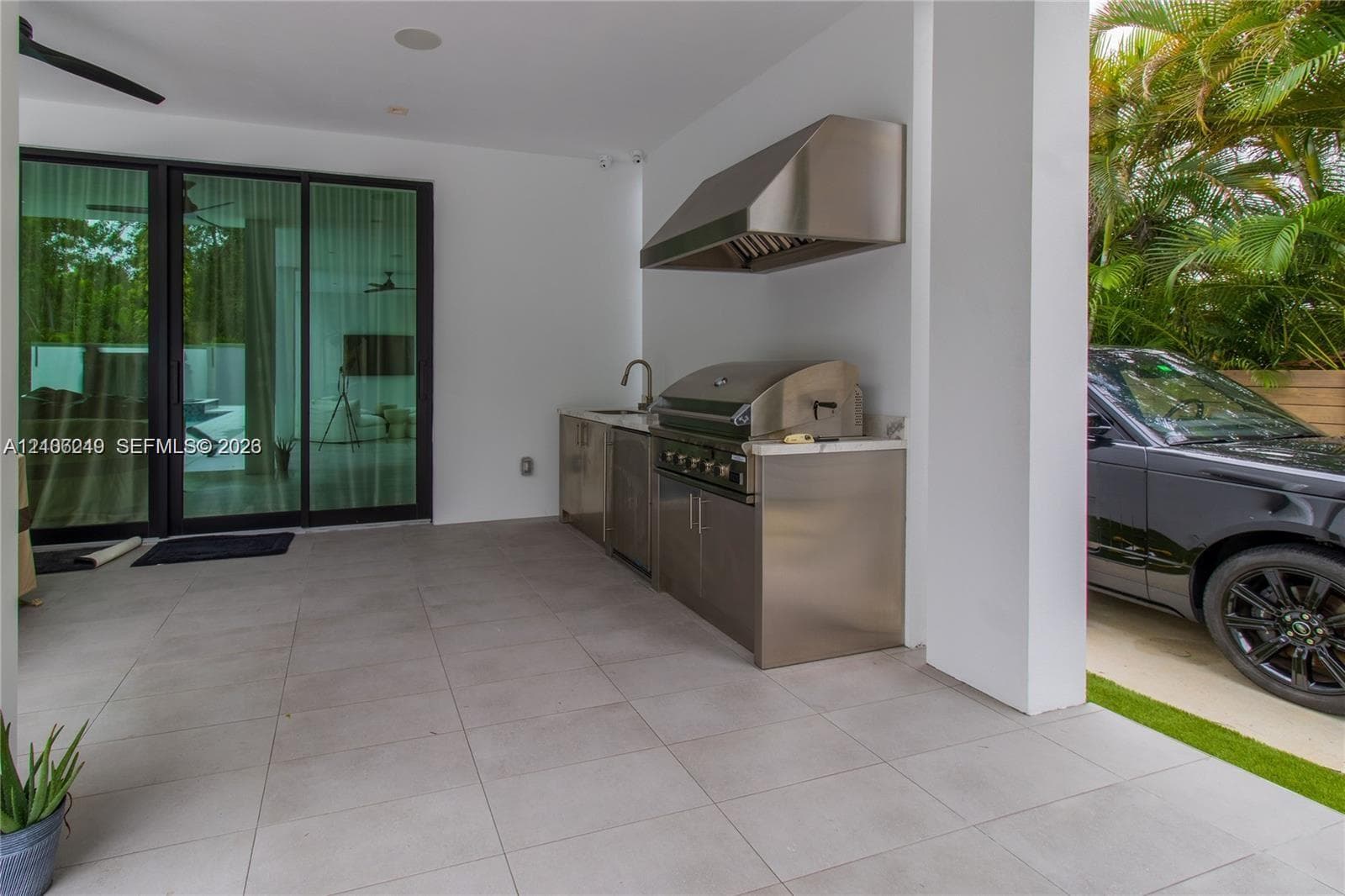 Covered outdoor kitchen featuring stainless steel cabinetry, built-in grill, professional-grade range hood, stone-surface countertop, and large-format porcelain tile flooring