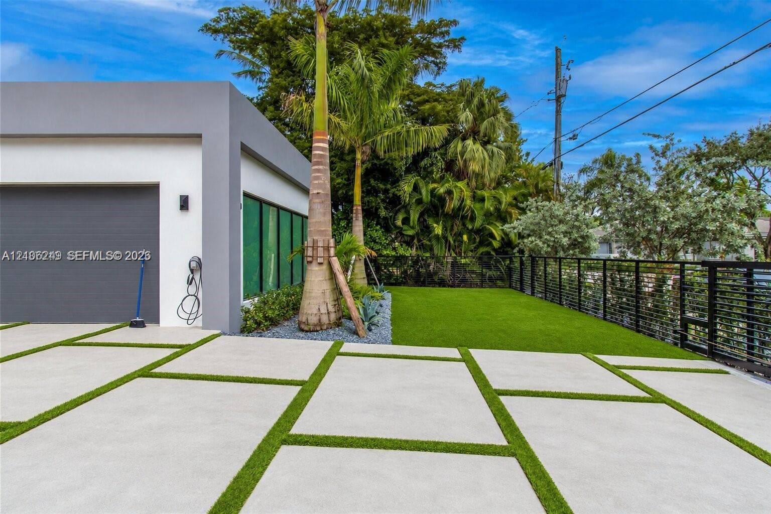 Modern exterior featuring a smooth gray and white facade, large-format concrete pavers with turf inlays, a single-car garage, and black metal perimeter fencing