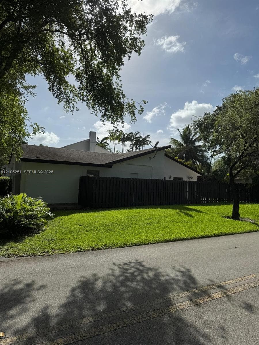 Stucco exterior residence featuring a dark wood-tone privacy fence, paved asphalt street access, and established mature trees