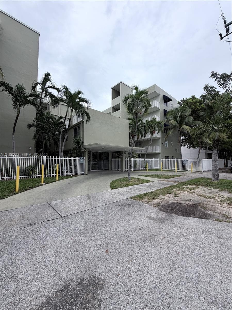 Multi-story building exterior featuring a covered entryway, concrete driveway, white metal fencing, and mature palm trees