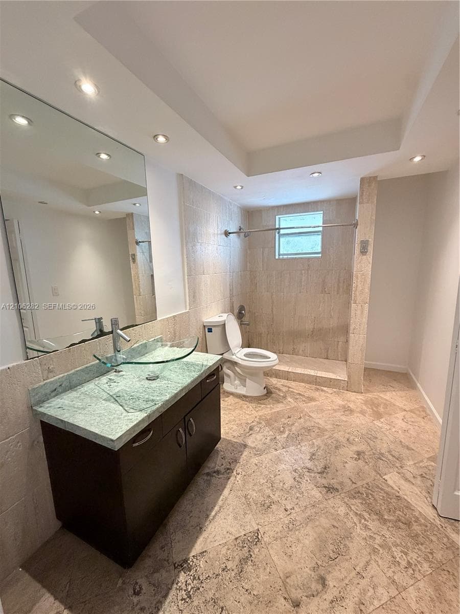 Bathroom featuring stone tile flooring and shower surround, dark wood-finish vanity with a stone countertop, glass vessel sink, and recessed ceiling lighting