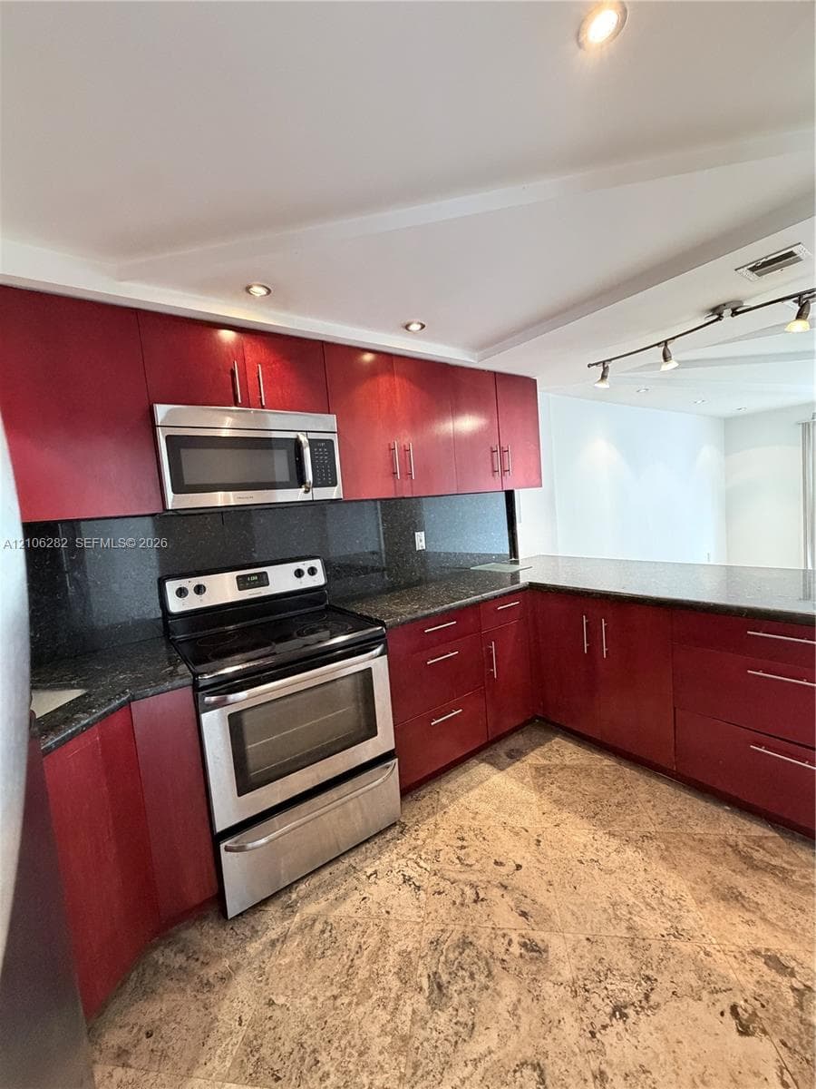 Kitchen featuring red-toned flat-panel cabinetry, dark solid-surface countertops, stainless steel appliance suite, stone flooring, and adjustable track lighting