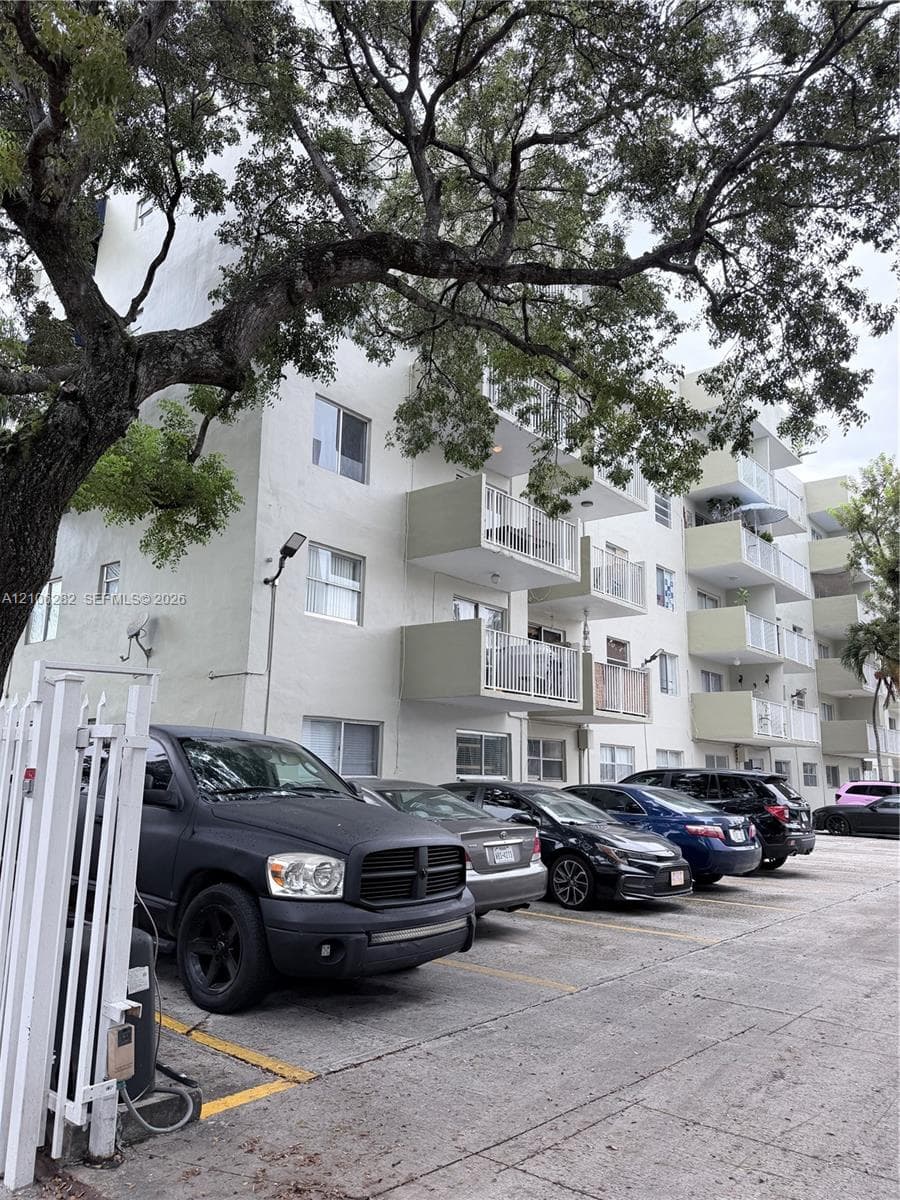 Multi-story exterior featuring a light-toned stucco façade, private balconies with metal railings, and paved surface parking