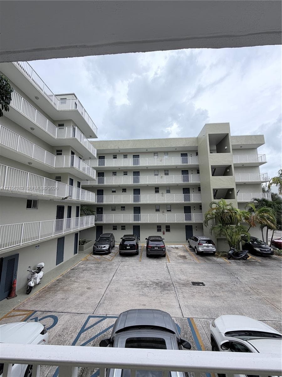 Multi-story exterior featuring concrete construction, white balcony railings, and paved surface parking