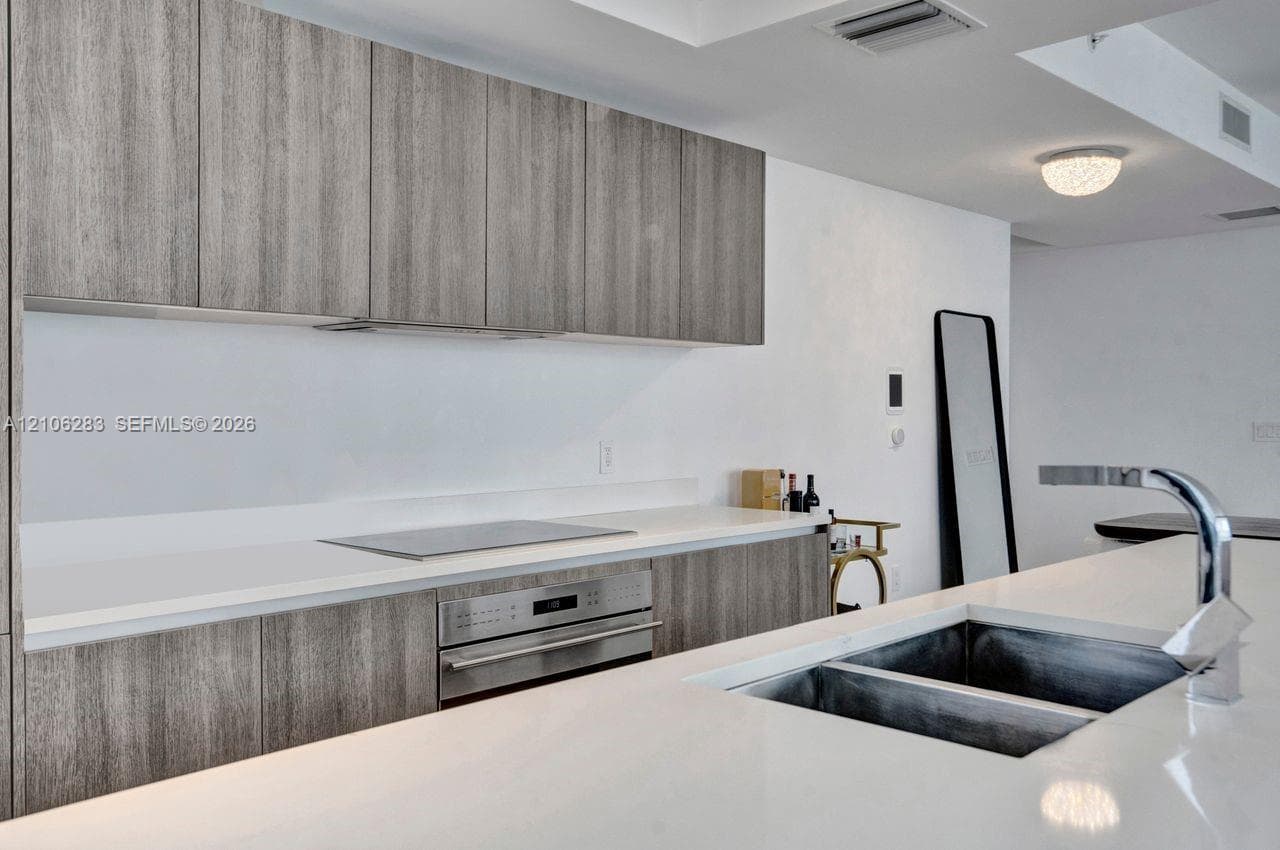 Kitchen featuring grey-toned wood-finish cabinetry, white composite countertops, a stainless steel wall oven, an integrated cooktop, and an undermount double-basin sink with a chrome faucet