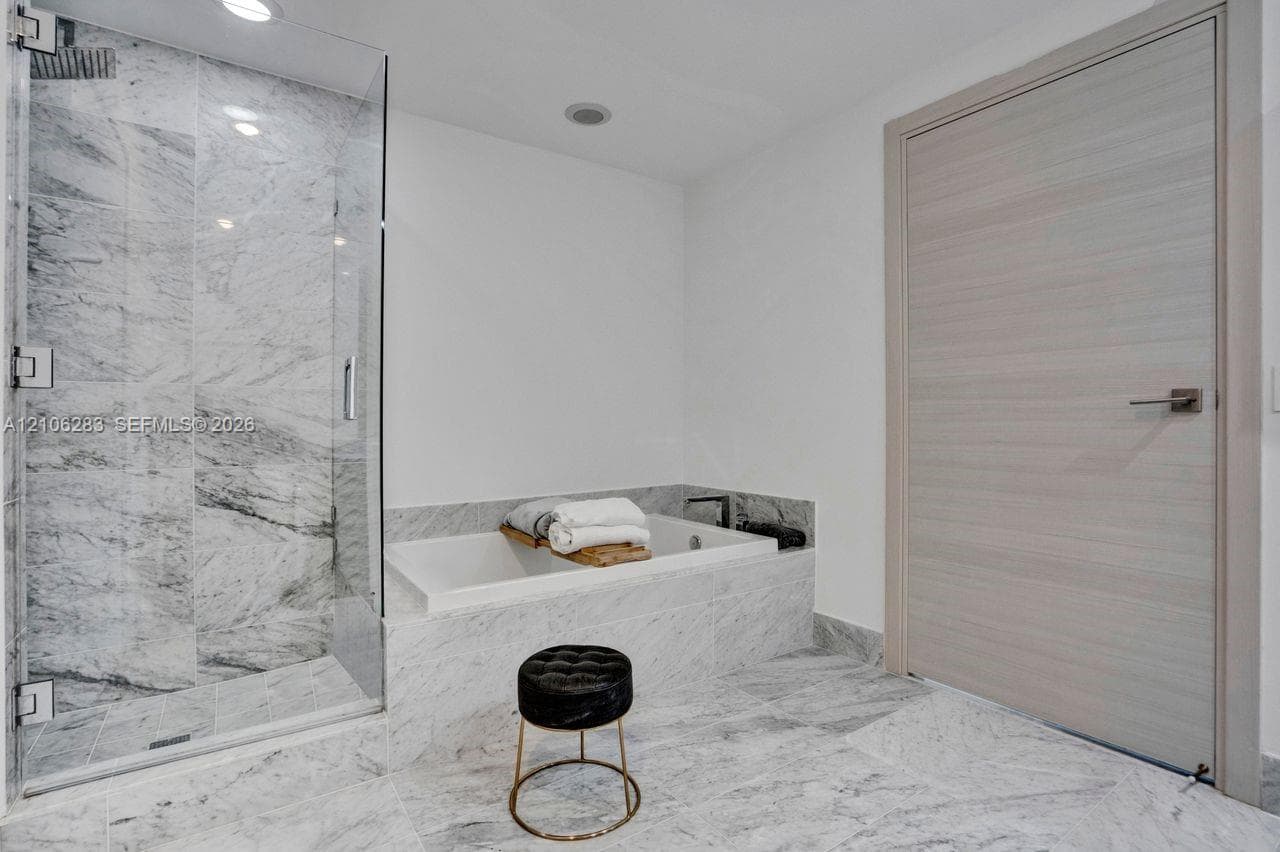 Frameless glass shower enclosure with stone tile surround, built-in soaking tub, stone tile flooring, and a light-toned wood-finish interior door