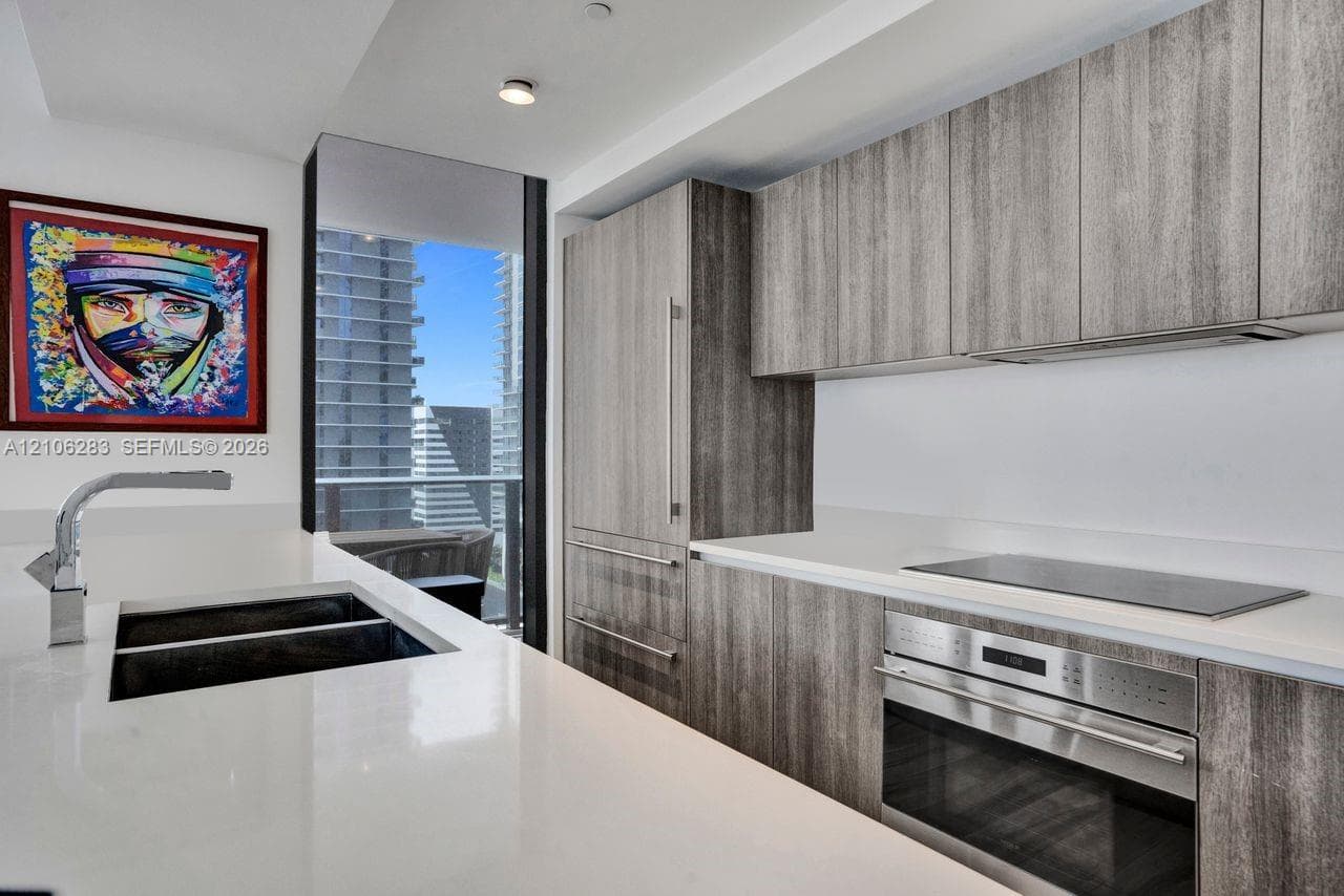 Kitchen featuring flat-panel wood-finish cabinetry, integrated stainless steel appliances, white solid-surface countertops, and a full-height floor-to-ceiling glass window