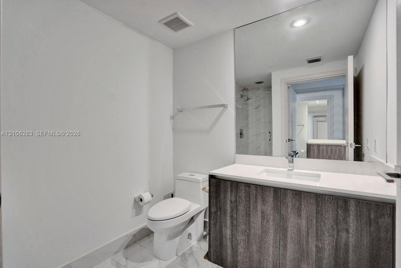 Modern vanity with dark wood-finish cabinetry, crisp white countertop, integrated rectangular sink, expansive wall-to-wall mirror, and stone-finish tile flooring
