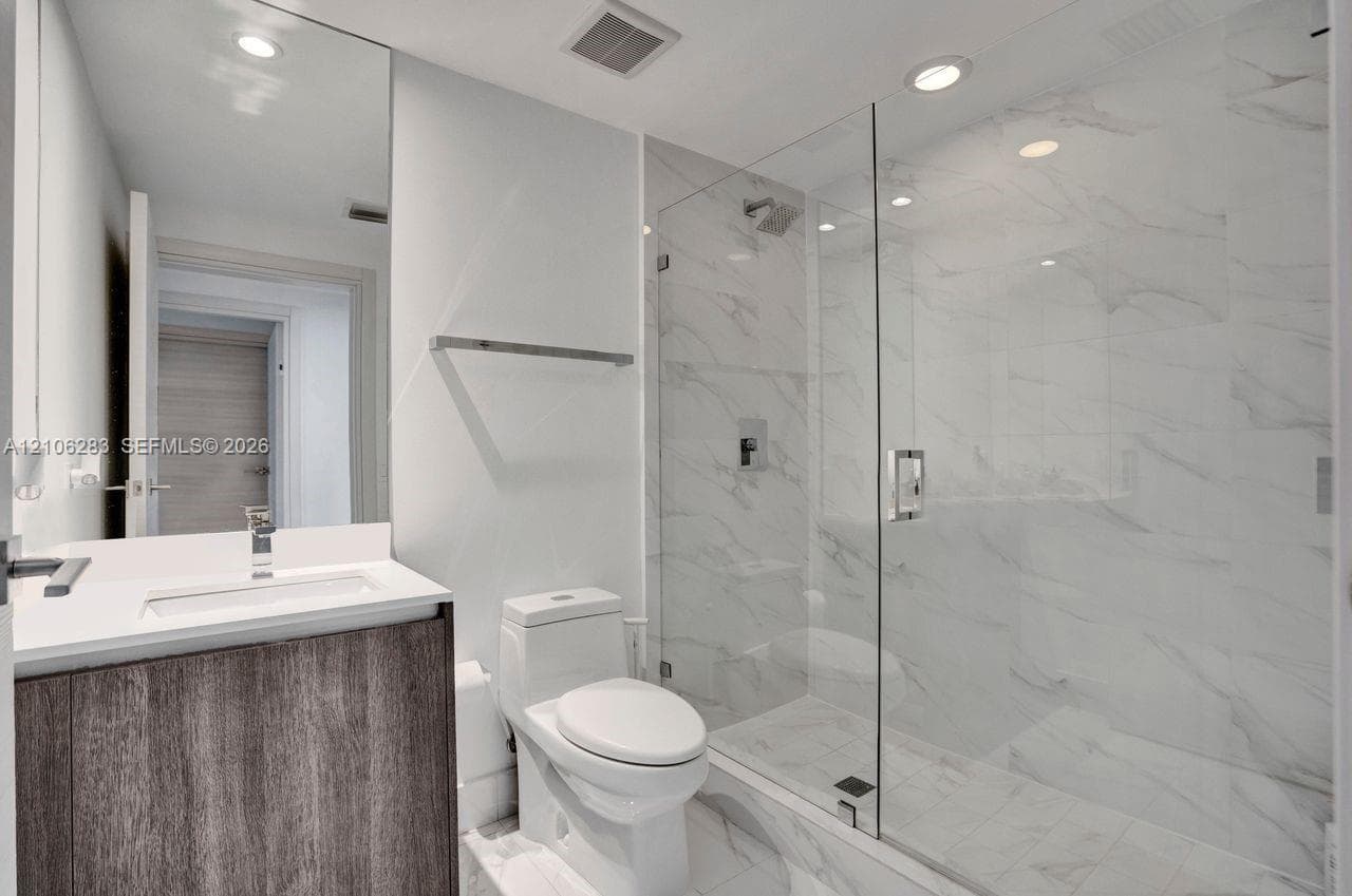Bathroom featuring a vanity with wood-finish cabinetry, a white countertop, a walk-in shower with a glass enclosure, floor-to-ceiling stone-style tile, and recessed lighting
