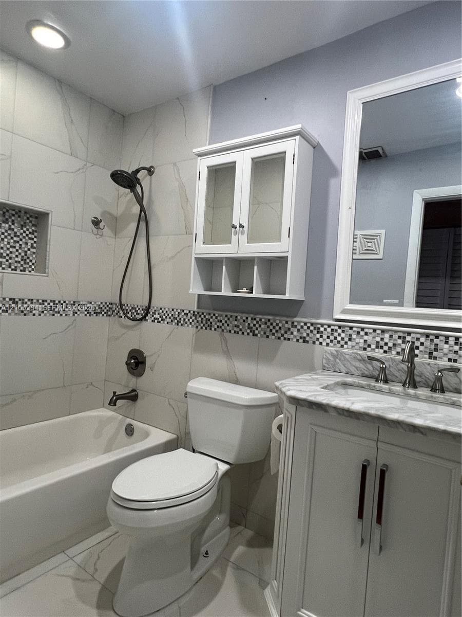 Bathroom featuring large-format light-toned tile walls, recessed shower niche with mosaic tile inlay, vanity with stone-finish countertop, mounted cabinetry, and framed mirror