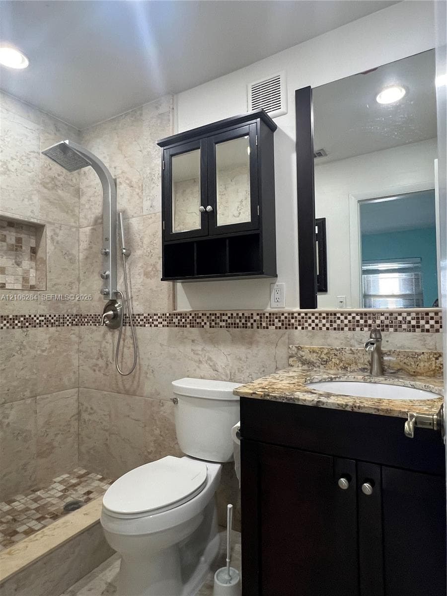 Full-tile shower stall with integrated recessed shelving, multi-head shower system, stone-finish vanity countertop with undermount sink, dark-finish cabinetry, and framed mirror