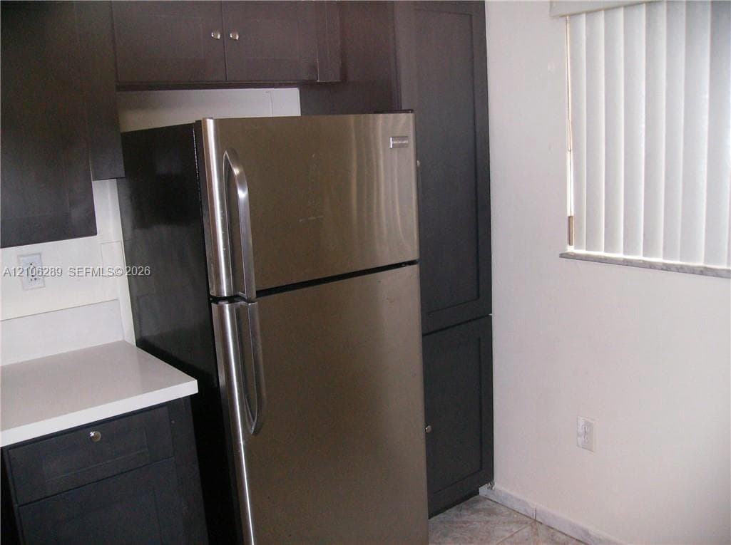 Dark-toned cabinetry, stainless steel refrigerator, white solid-surface countertops, and vertical window blinds